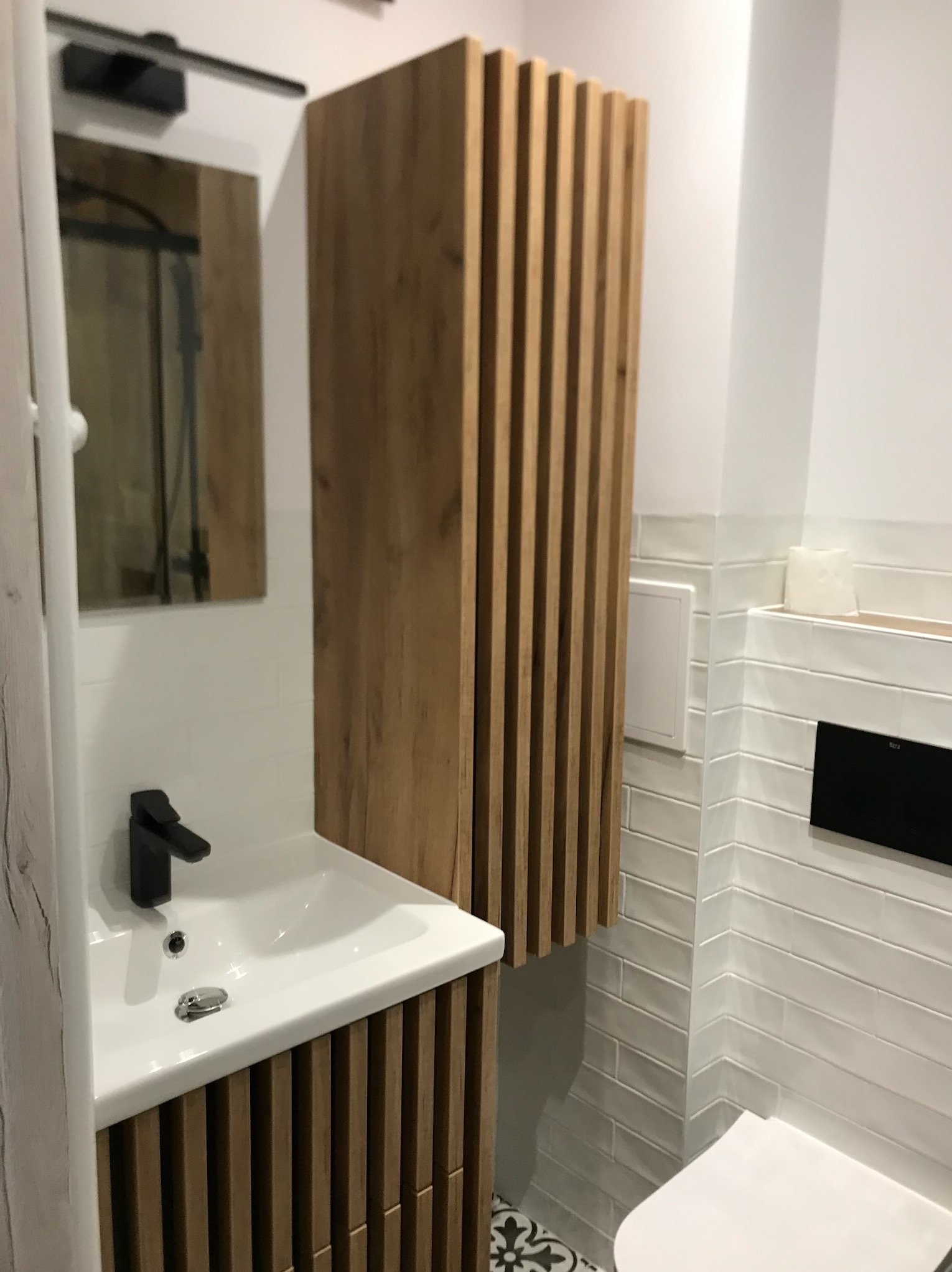 Nowoczesna łazienka: biała umywalka z szafką z drewnianymi lamelami, czarna bateria, lustro i białe płytki ułożone w jodełkę. Dekoracyjne płytki na podłodze.