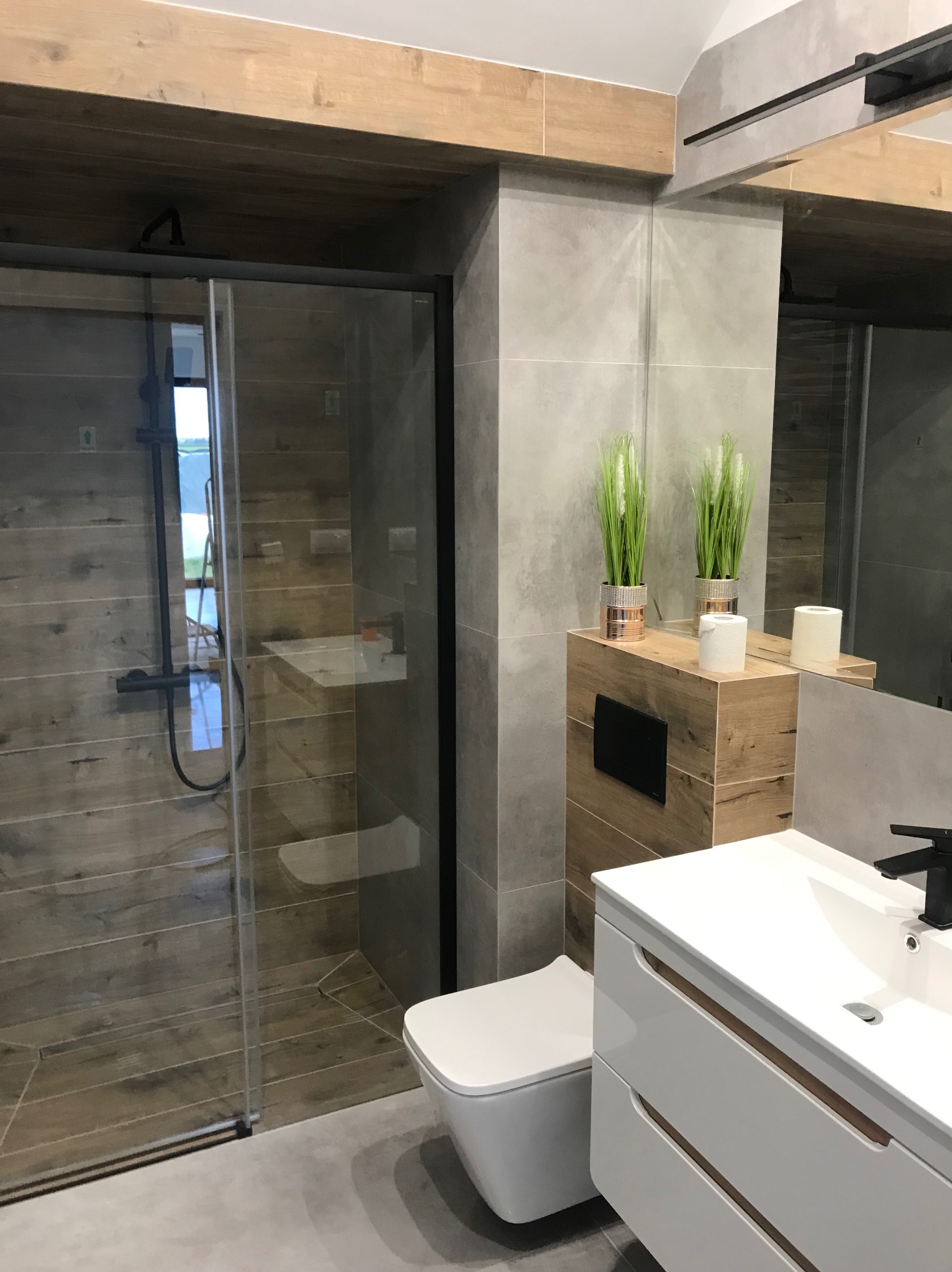 Nowoczesna łazienka po remoncie: szklana kabina prysznicowa z czarnymi profilami, drewnopodobne płytki, biała umywalka z szafką i akcenty roślinne.