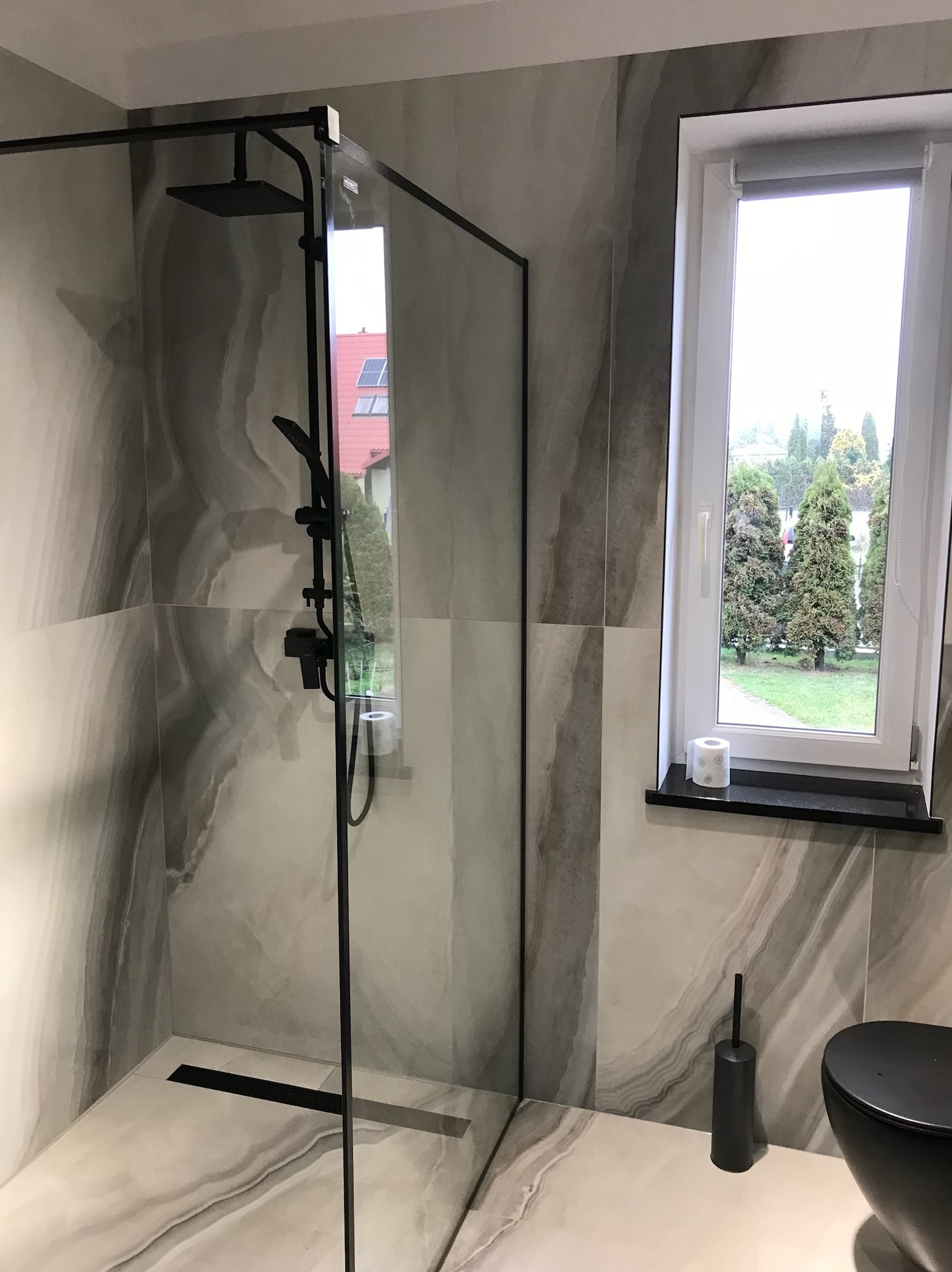 Nowoczesna łazienka z kabiną typu walk-in, czarną armaturą i płytkami imitującymi marmur. Widoczne okno z widokiem na zieleń.