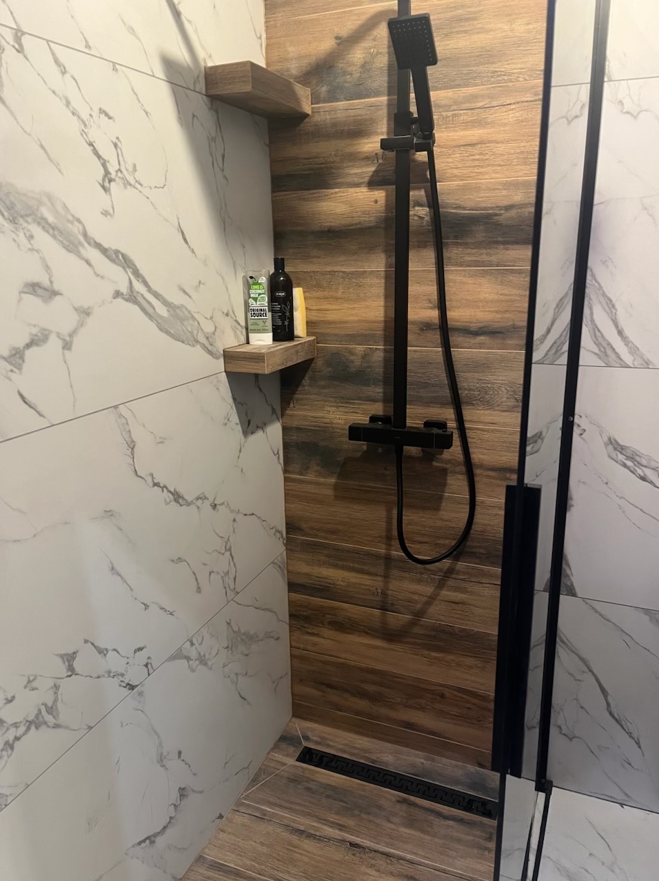 Nowoczesna kabina prysznicowa z drewnopodobnymi i marmurowymi płytkami, czarną armaturą i półkami z kosmetykami. Widoczny odpływ liniowy.