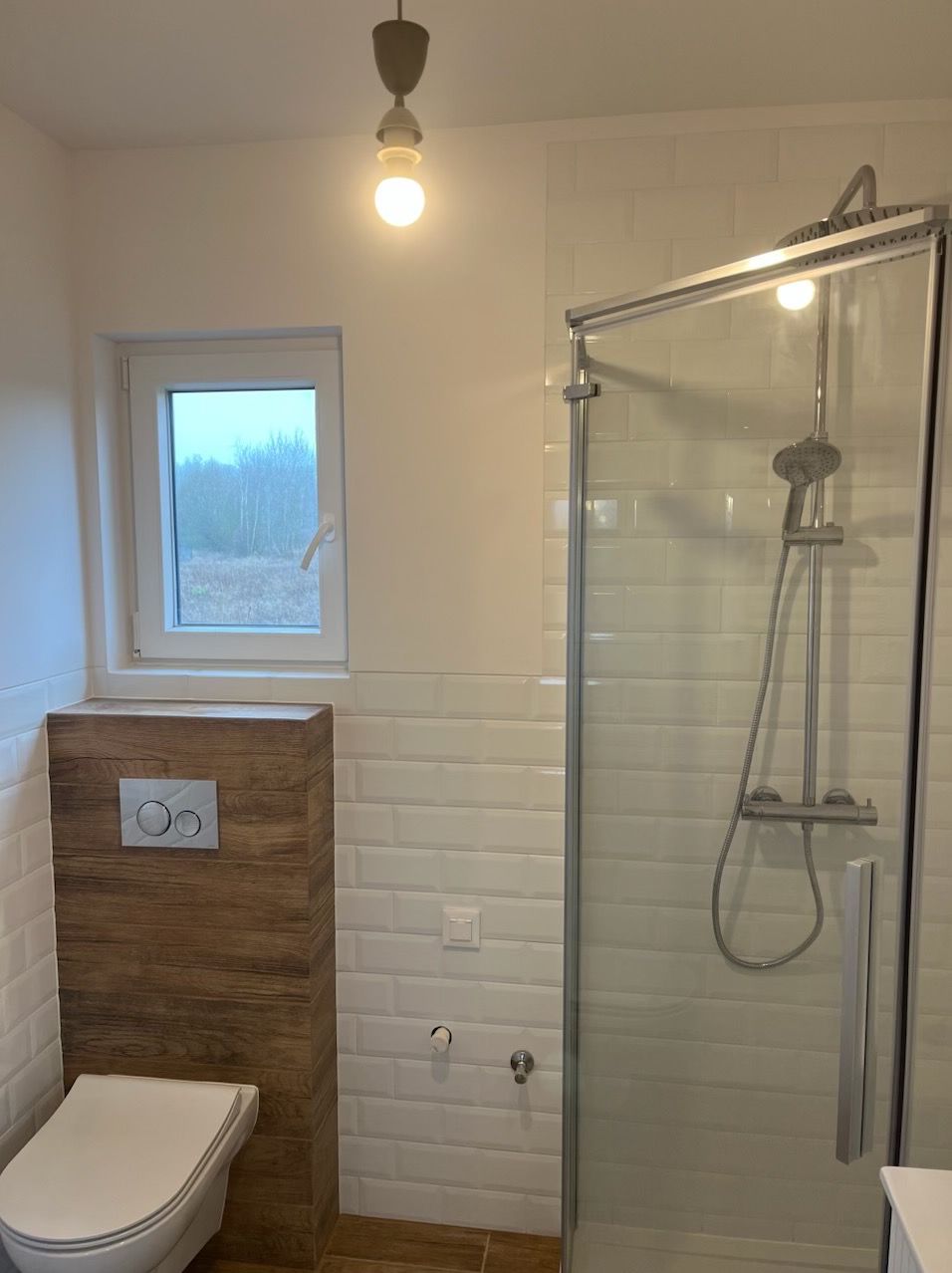 Nowoczesna łazienka z kabiną prysznicową i zabudowaną toaletą. Białe płytki imitujące cegłę, drewnopodobna obudowa toalety, okno z widokiem na zewnątrz.