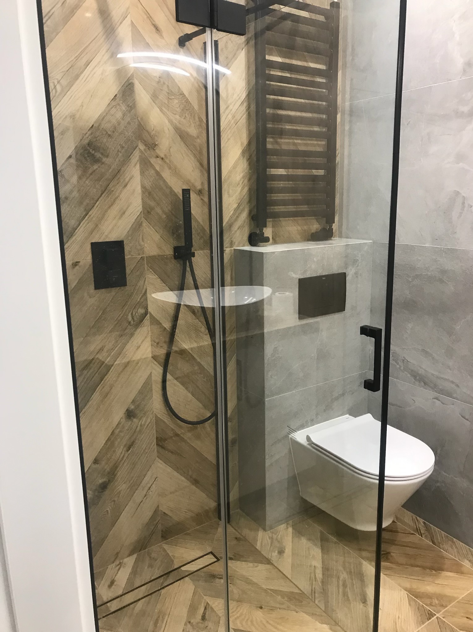Nowoczesna łazienka z kabiną prysznicową typu walk-in. Uwagę przykuwają drewnopodobne płytki ułożone w jodełkę oraz czarne akcenty armatury i grzejnika.