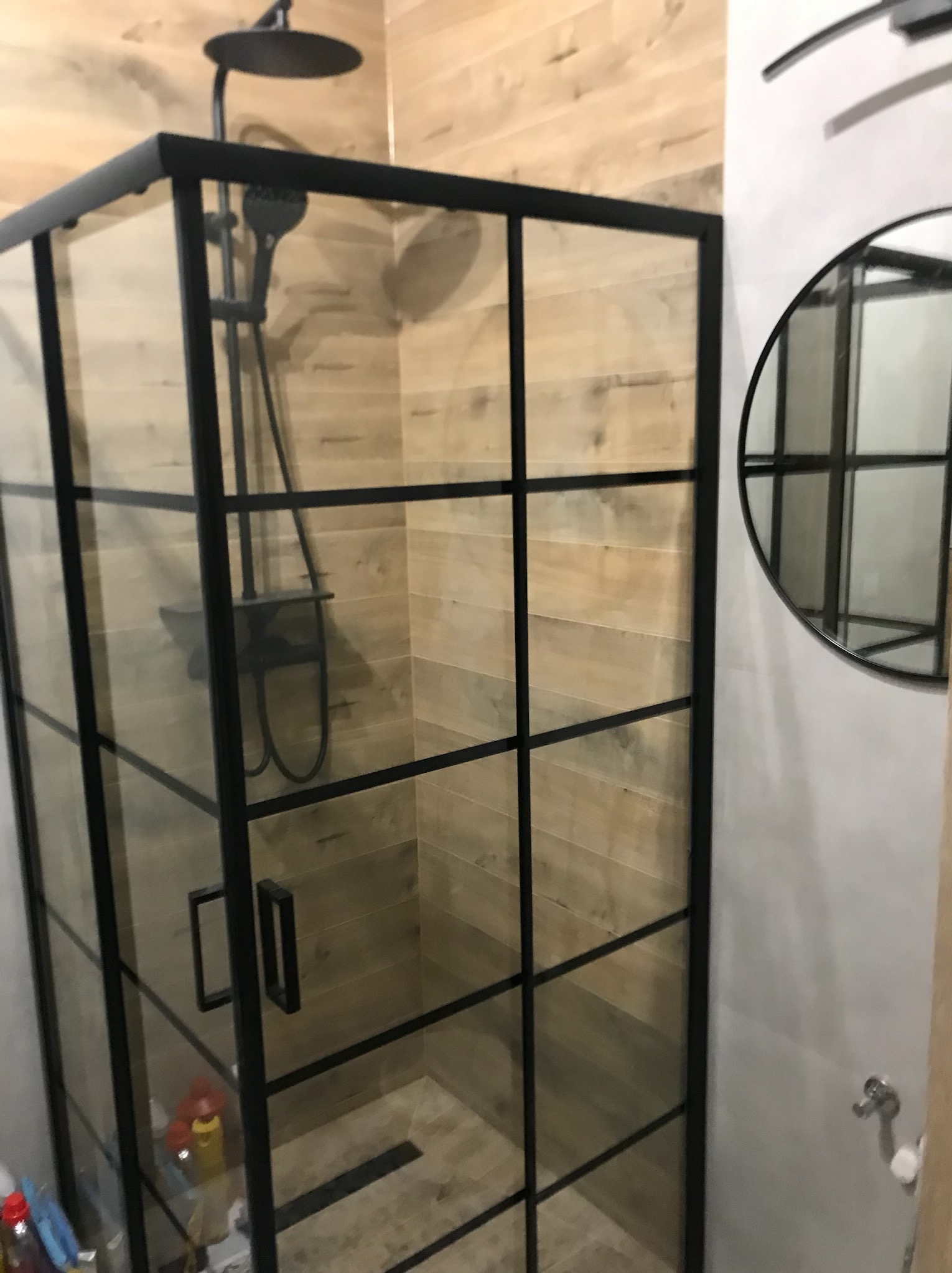 Kabina prysznicowa z czarnymi profilami i armaturą, płytki imitujące drewno na ścianach, okrągłe lustro. Nowoczesna aranżacja łazienki.