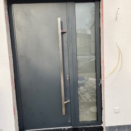 TEXX-OKNA - Montaż szarych drzwi zewnętrznych z długim, pionowym uchwytem i bocznym oknem. Widoczne niedokończone prace wykończeniowe wokół ramy i wystające przewody elektryczne.