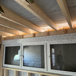 TEXX-OKNA - Montaż trzech białych okien w nowo budowanym domu, widoczne drewniane belki stropowe i konstrukcja ścian.