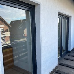 TEXX-OKNA - Dwa przesuwne okna balkonowe z ciemnymi ramami na tle białej, chropowatej ściany zewnętrznej. W odbiciu szyb widoczne budynki mieszkalne i niebo.