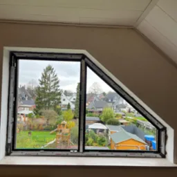 Narożne okno PCV z czarną ramą, częściowo otwarte, widok na ogród z domkami i placem zabaw w tle, na parapecie grzejnik.