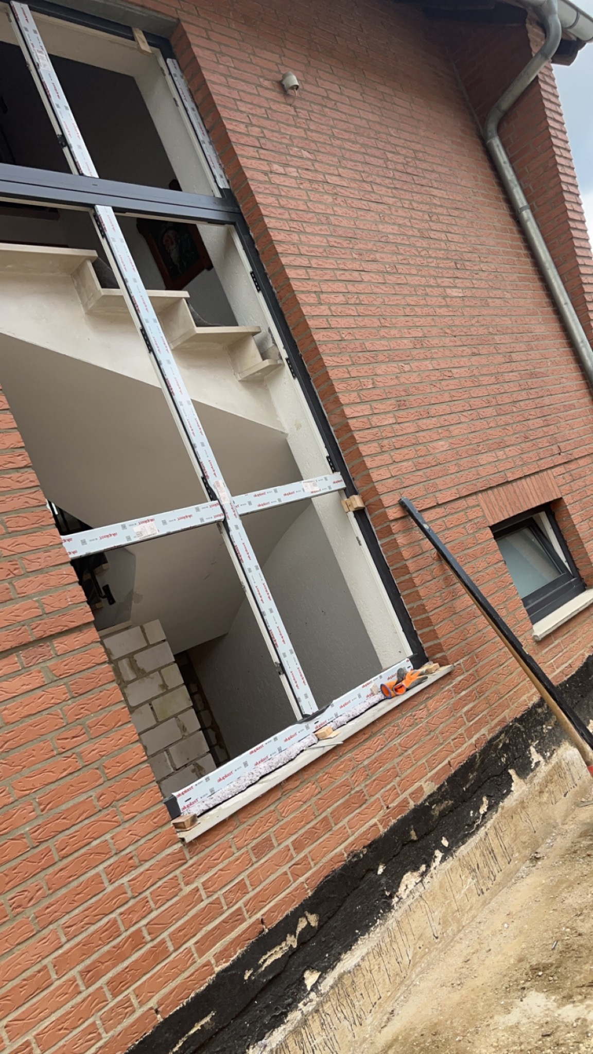 Montaż okna w surowym stanie budynku z cegły, widoczne narzędzia i elementy mocujące w otworze okiennym, schody w tle.