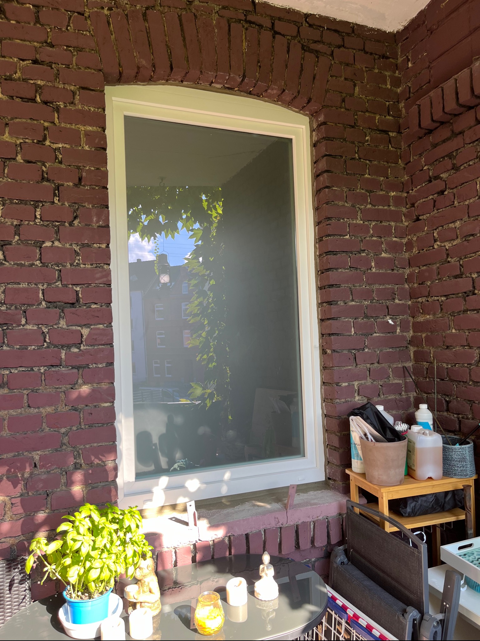 Białe okno w czerwonej ceglanej ścianie, widok na zewnątrz z zielonymi liśćmi i budynkiem w tle, fragment balkonu z roślinami i stolikiem.