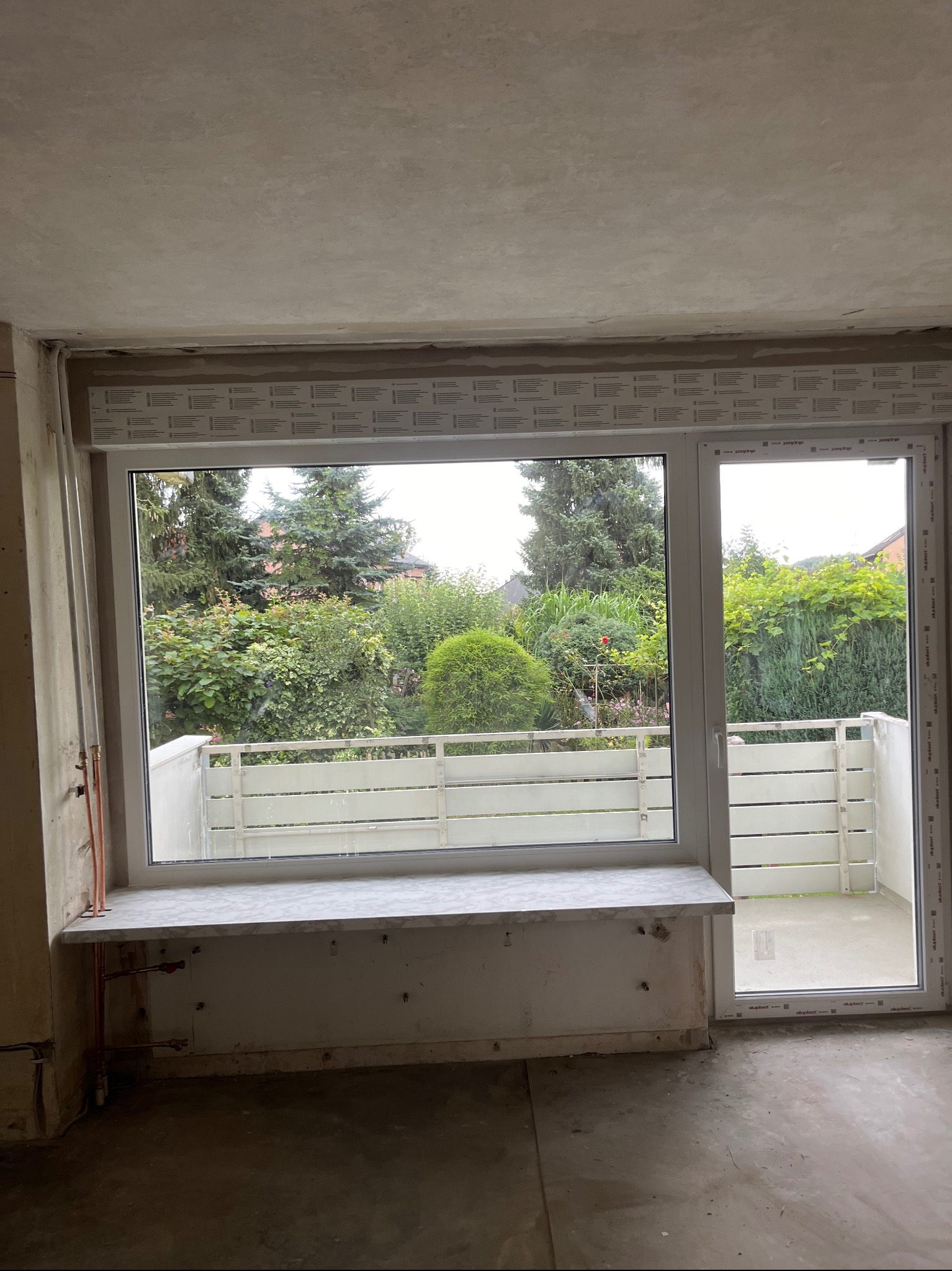 Nowe okno PCV z roletą zewnętrzną, widok na balkon i zieleń z wnętrza w trakcie remontu.