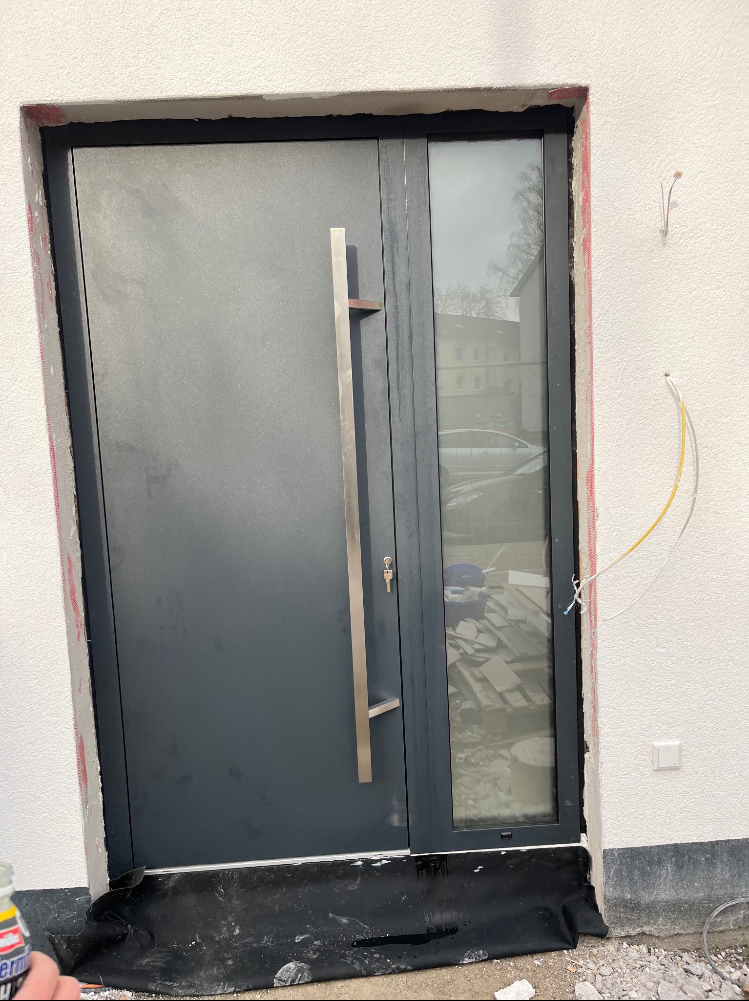 Montaż szarych drzwi zewnętrznych z długim, pionowym uchwytem i bocznym oknem. Widoczne niedokończone prace wykończeniowe wokół ramy i wystające przewody elektryczne.
