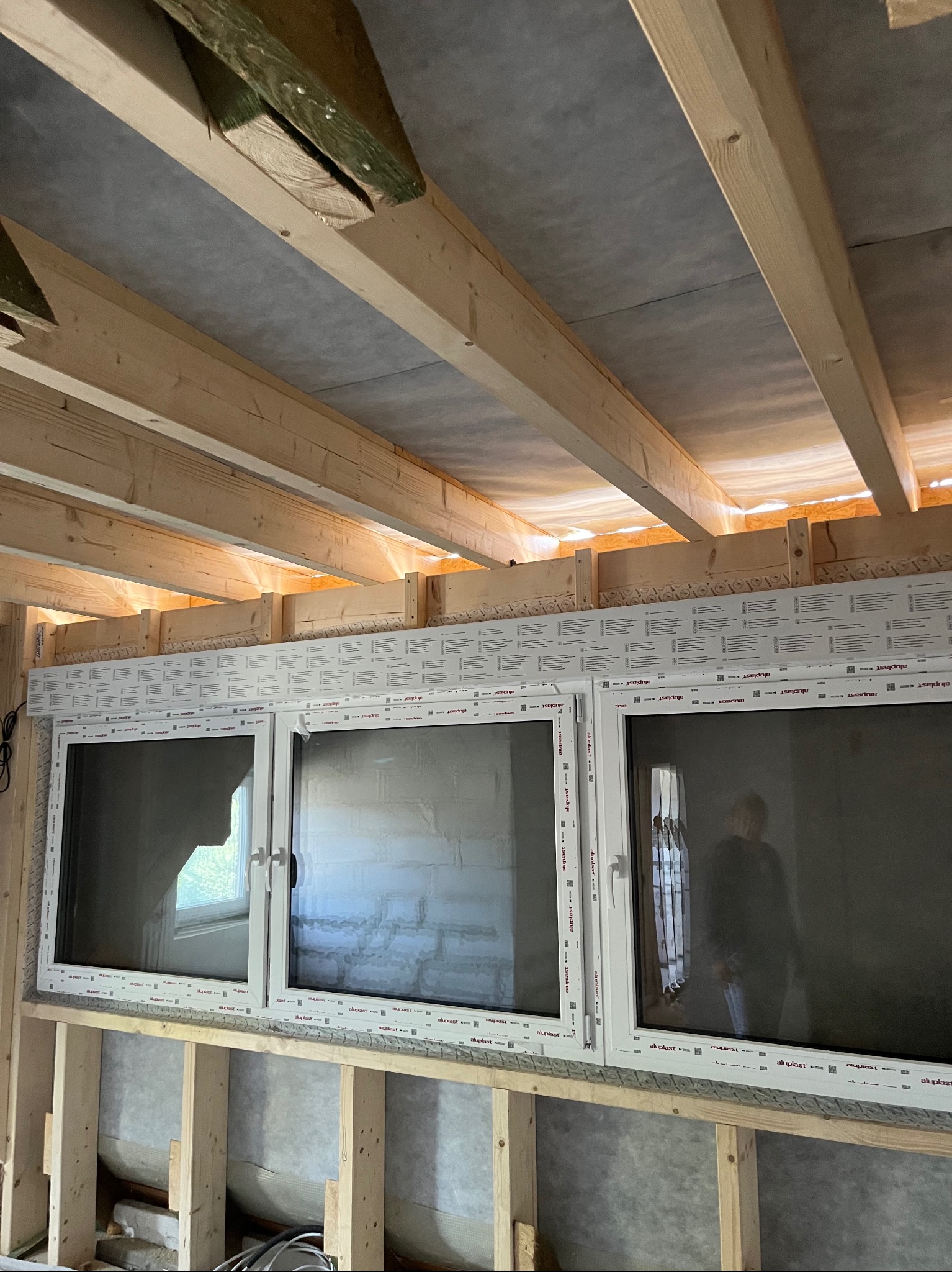 Montaż trzech białych okien w nowo budowanym domu, widoczne drewniane belki stropowe i konstrukcja ścian.
