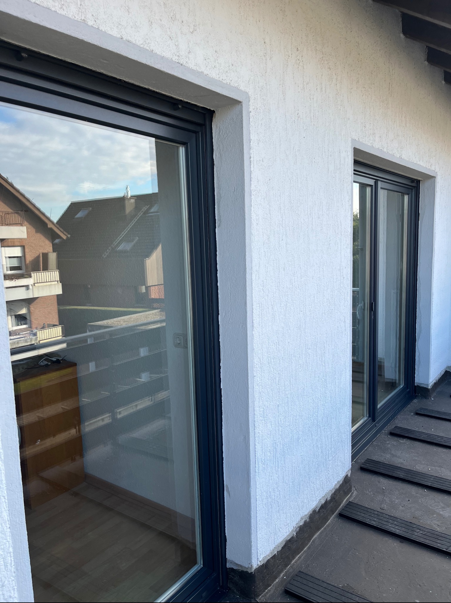 Dwa przesuwne okna balkonowe z ciemnymi ramami na tle białej, chropowatej ściany zewnętrznej. W odbiciu szyb widoczne budynki mieszkalne i niebo.