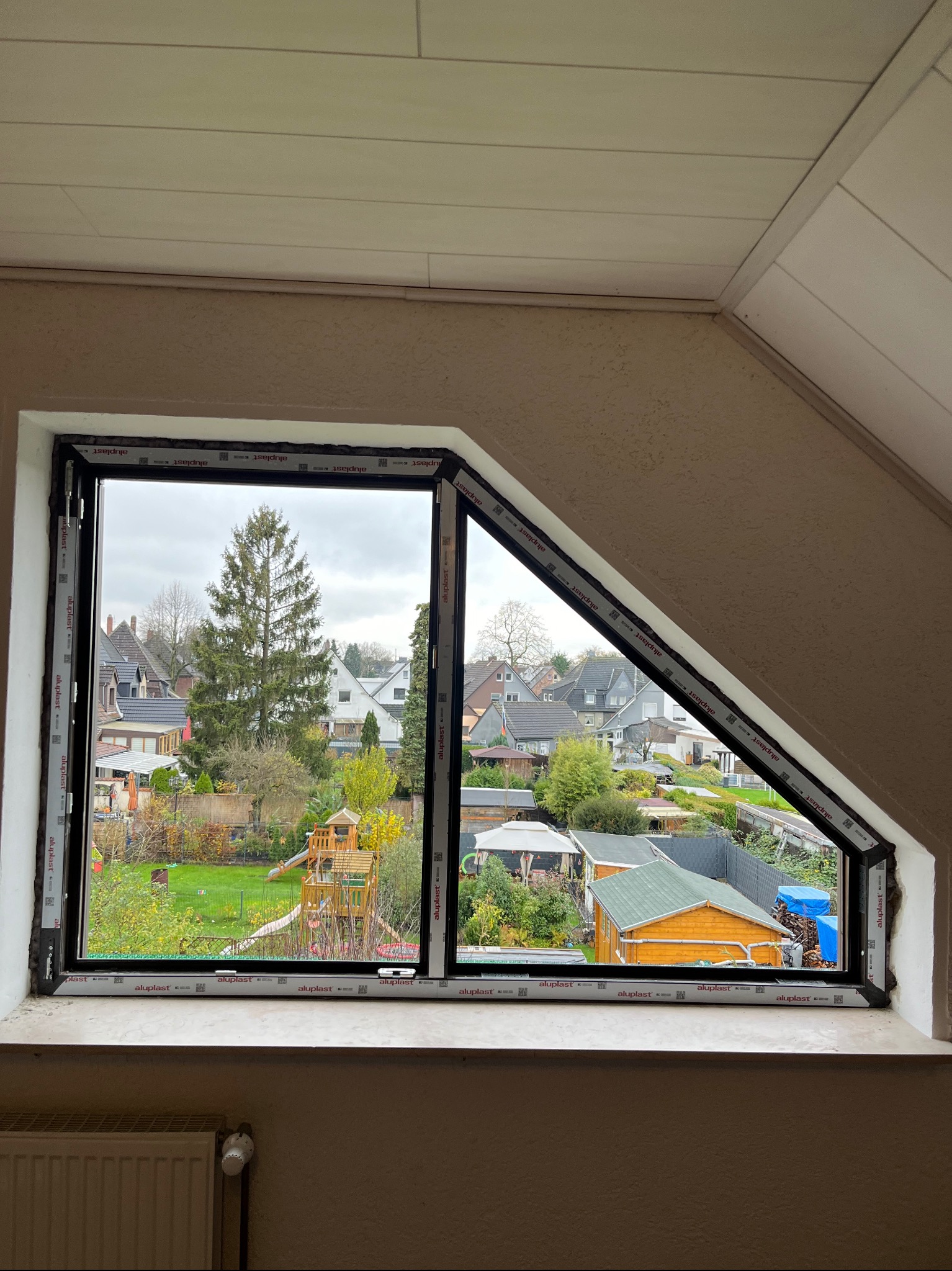 Narożne okno PCV z czarną ramą, częściowo otwarte, widok na ogród z domkami i placem zabaw w tle, na parapecie grzejnik.