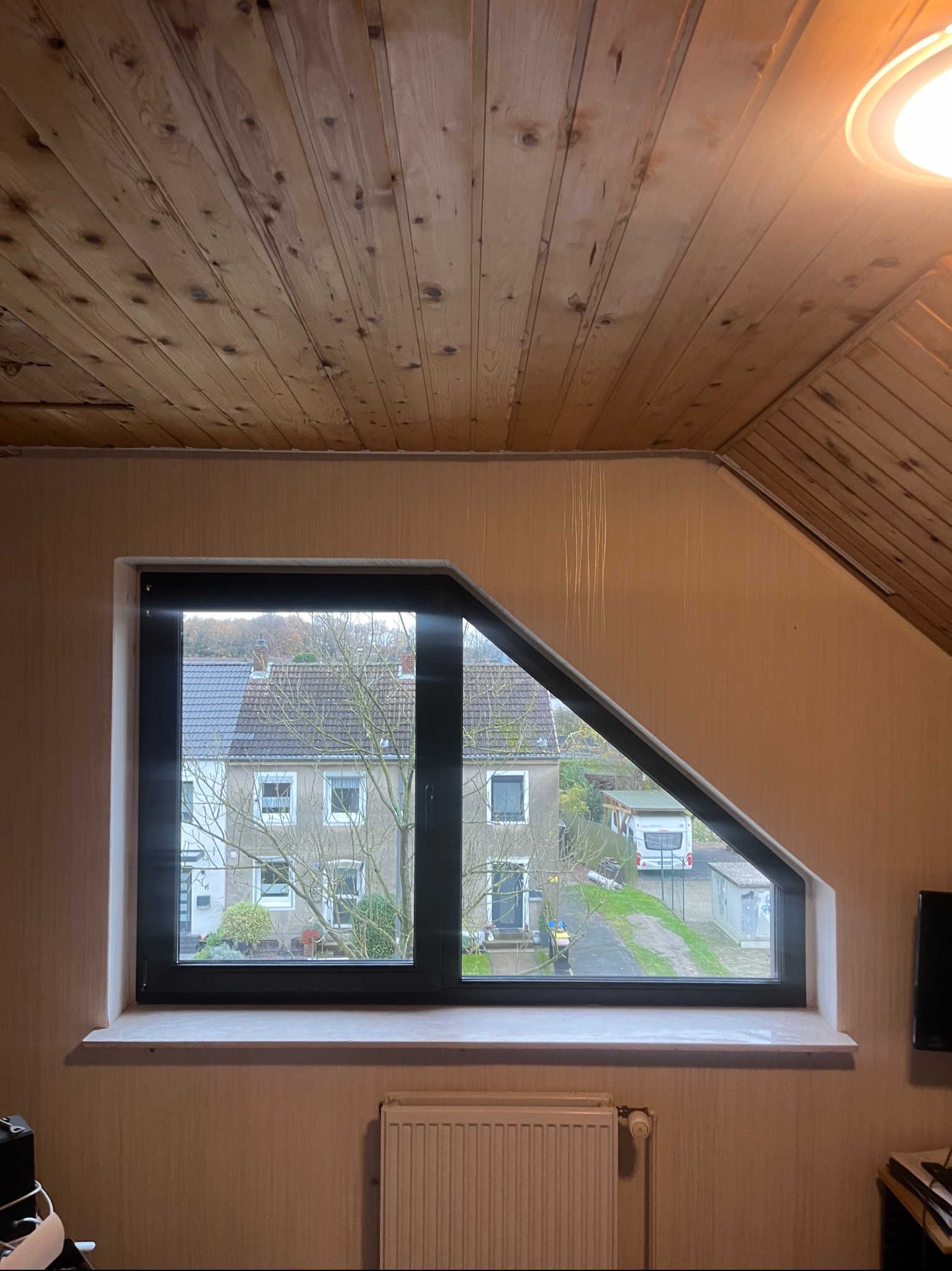 Nietypowe okno o czarnej ramie, z ukośną górną krawędzią, w pokoju z drewnianą boazerią na suficie, widok na domy i drzewa na zewnątrz.