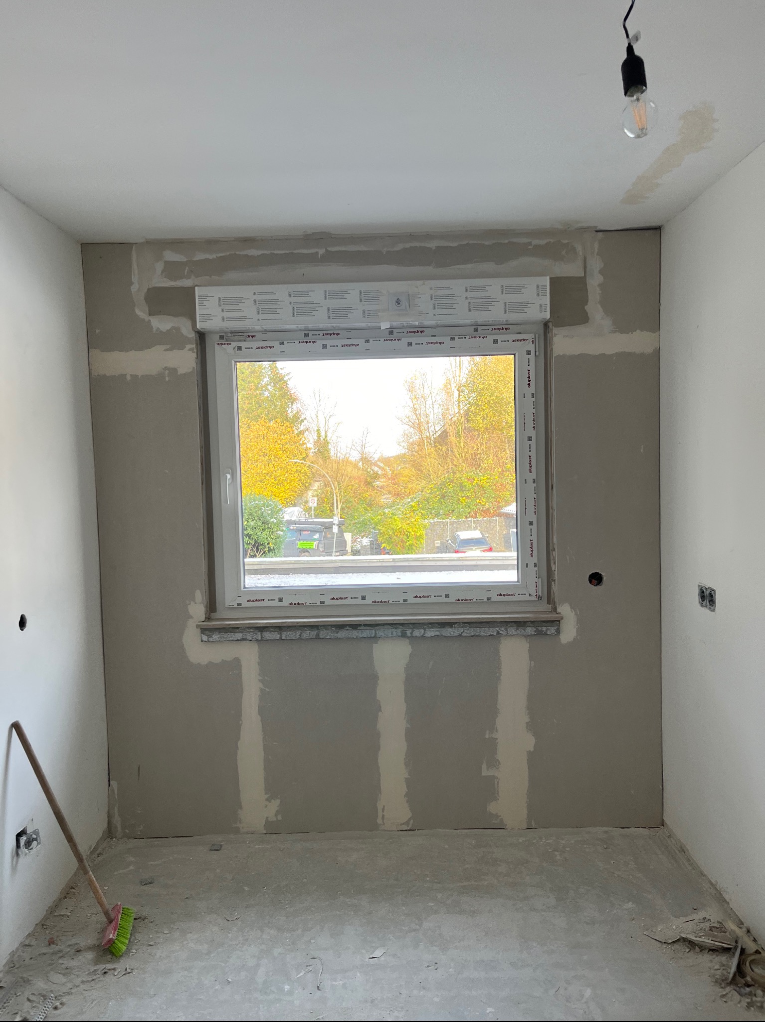 Pokój w trakcie remontu z zamontowanym oknem PCV z częściowo opuszczoną roletą zewnętrzną, ściany w stanie surowym, widoczne ślady gładzi, na podłodze resztki gruzu i miotła