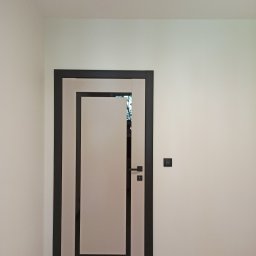 Yurii Radoman - Nowoczesne drzwi wewnętrzne z czarną ramą i jasnym wypełnieniem, zamontowane w białym pomieszczeniu z drewnianą podłogą. Widoczny włącznik światła.