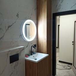 Yurii Radoman - Nowoczesna łazienka z marmurową ścianą, okrągłym lustrem z podświetleniem LED, drewnianą szafką pod umywalką i czarną armaturą. Widok na otwarte drzwi.