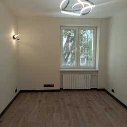 Yurii Radoman - Jasne wnętrze po remoncie: drewnopodobne panele, białe ściany, okno z widokiem na zieleń, nowoczesna lampa sufitowa i ścienna. Minimalistyczny design.