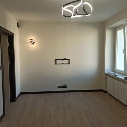 Yurii Radoman - Jasne wnętrze po remoncie: parkiet, świeżo pomalowane ściany z nowoczesnym oświetleniem, listwy przypodłogowe i przygotowane miejsce na telewizor.