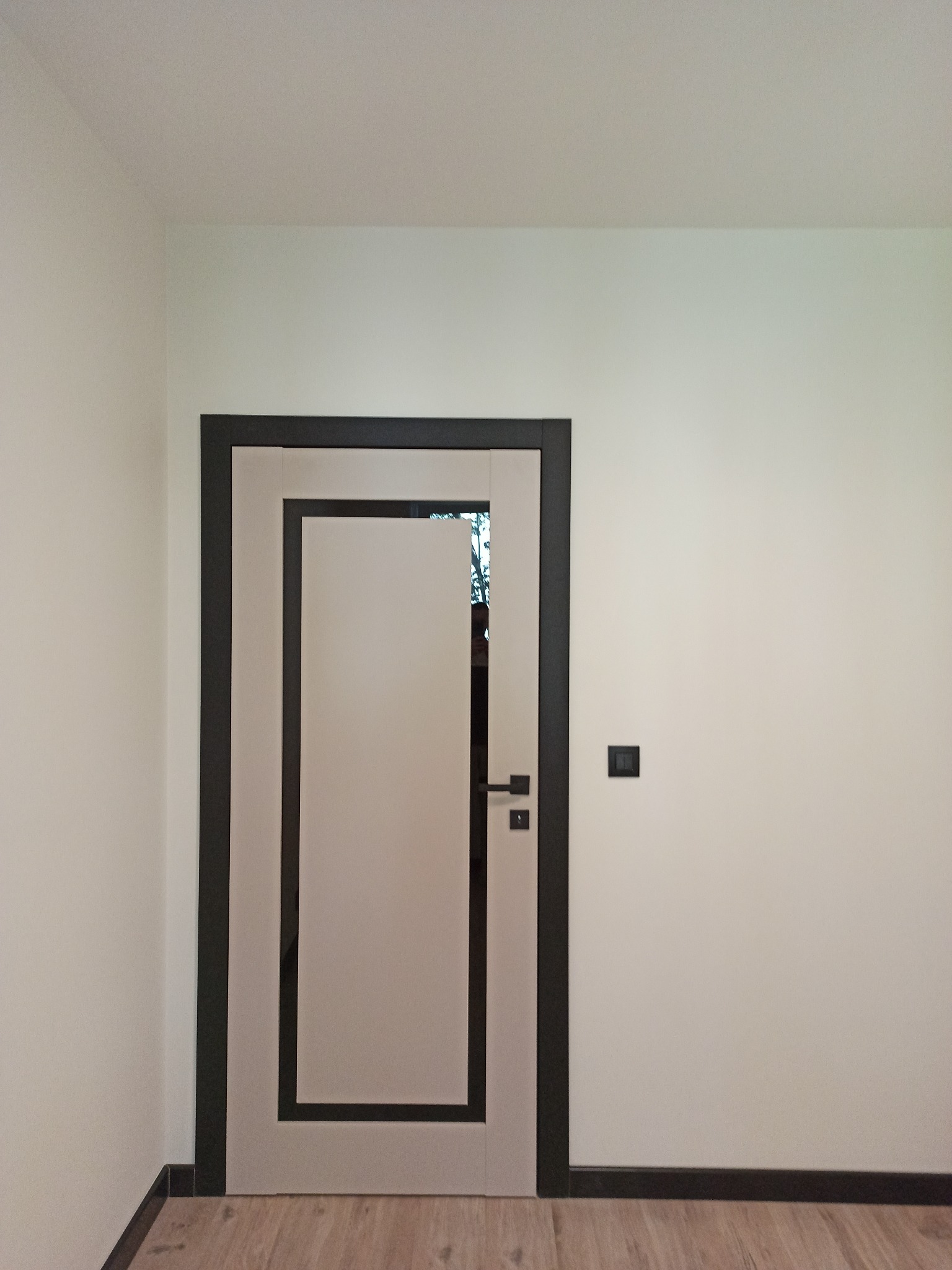 Nowoczesne drzwi wewnętrzne z czarną ramą i jasnym wypełnieniem, zamontowane w białym pomieszczeniu z drewnianą podłogą. Widoczny włącznik światła.