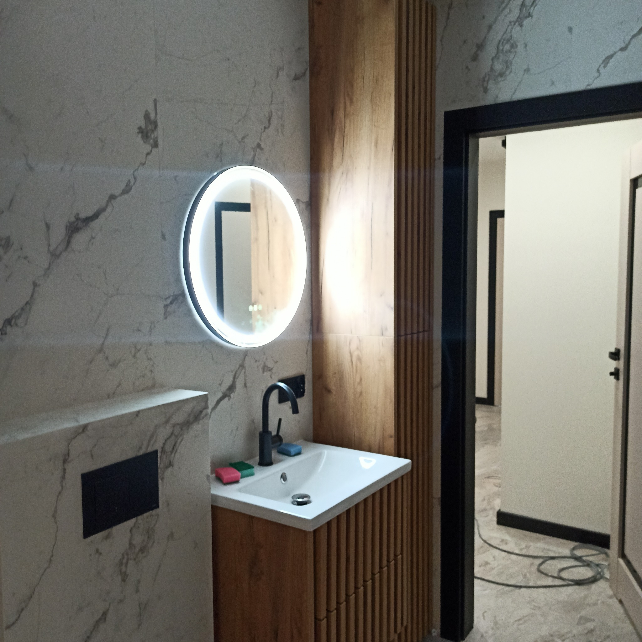 Nowoczesna łazienka z marmurową ścianą, okrągłym lustrem z podświetleniem LED, drewnianą szafką pod umywalką i czarną armaturą. Widok na otwarte drzwi.