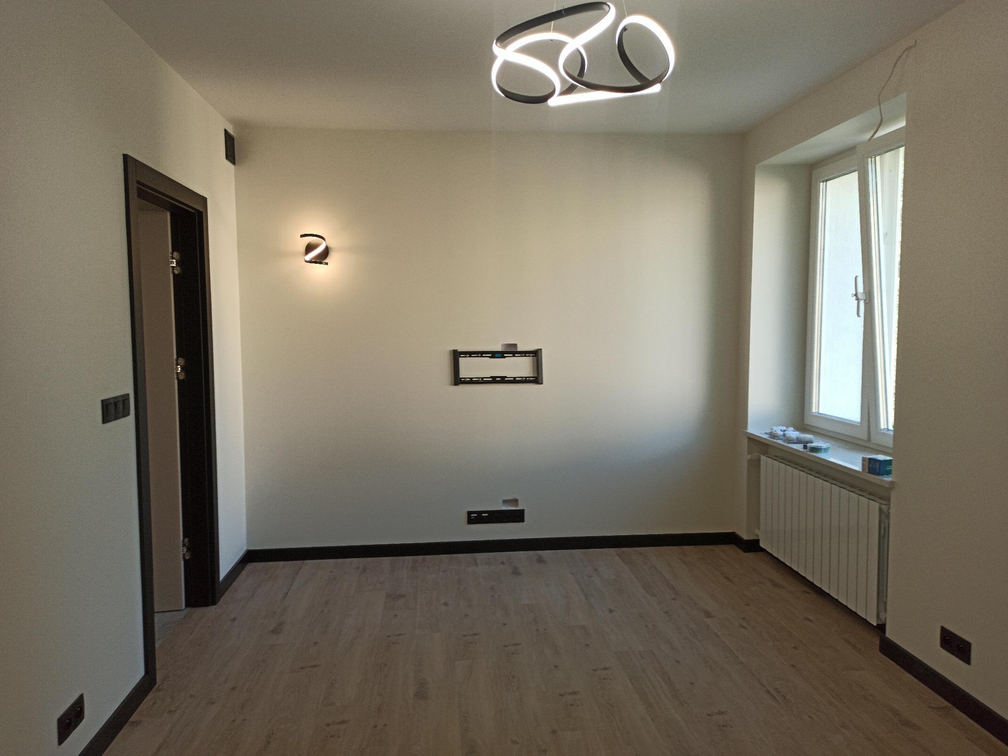 Jasne wnętrze po remoncie: parkiet, świeżo pomalowane ściany z nowoczesnym oświetleniem, listwy przypodłogowe i przygotowane miejsce na telewizor.