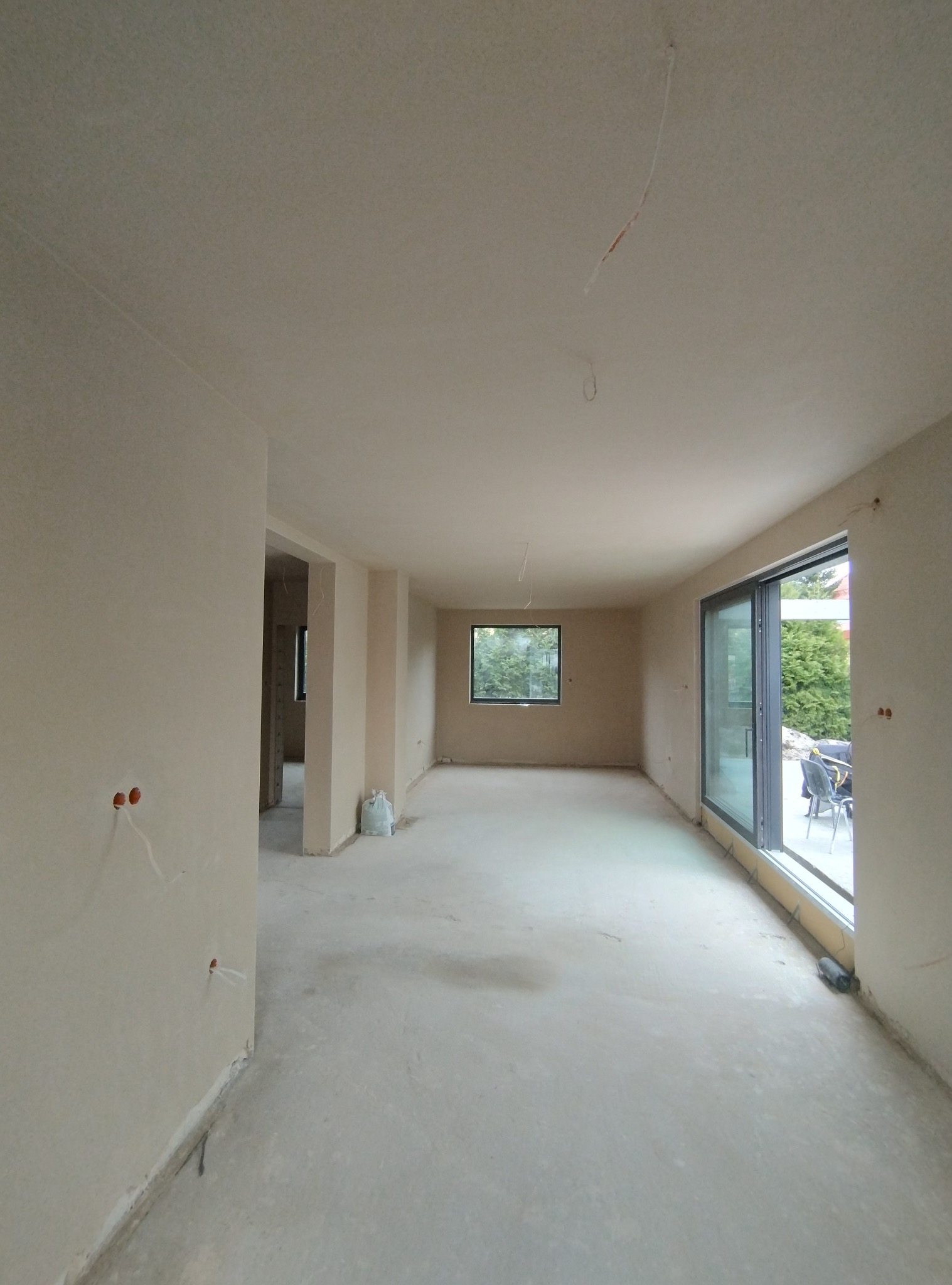 Wnętrze domu w trakcie prac wykończeniowych, ściany i sufit po nałożeniu gładzi, widoczne wystające kable i worki z materiałami budowlanymi.