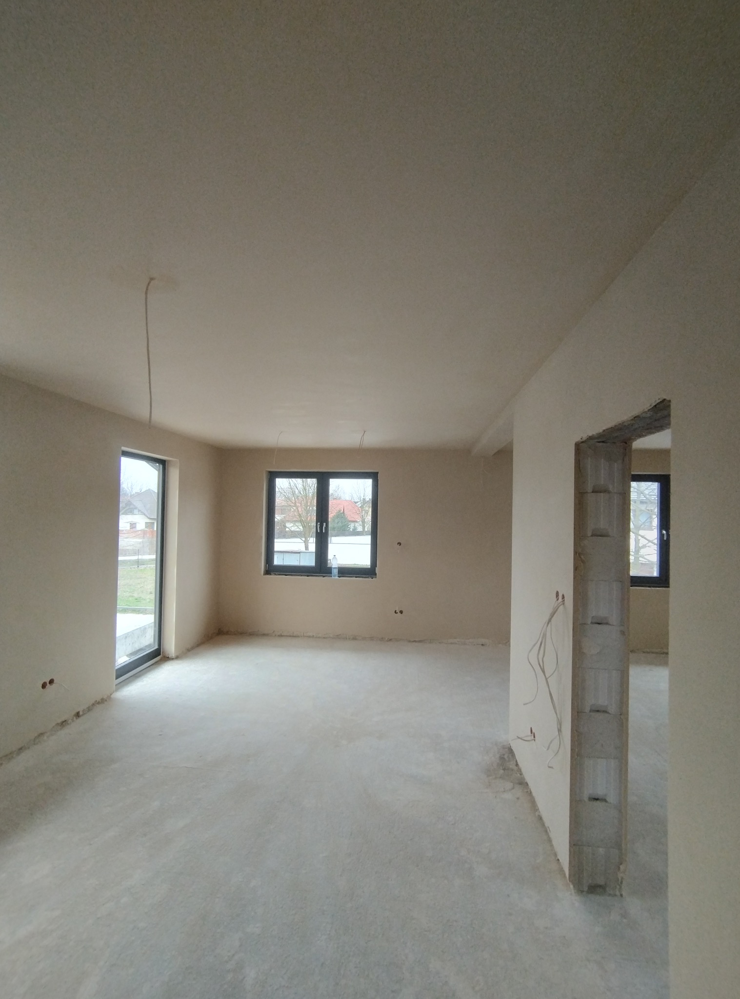 Surowe wnętrze domu po nałożeniu gładzi, widoczne okna z czarnymi ramami i wystające przewody elektryczne, stan deweloperski.