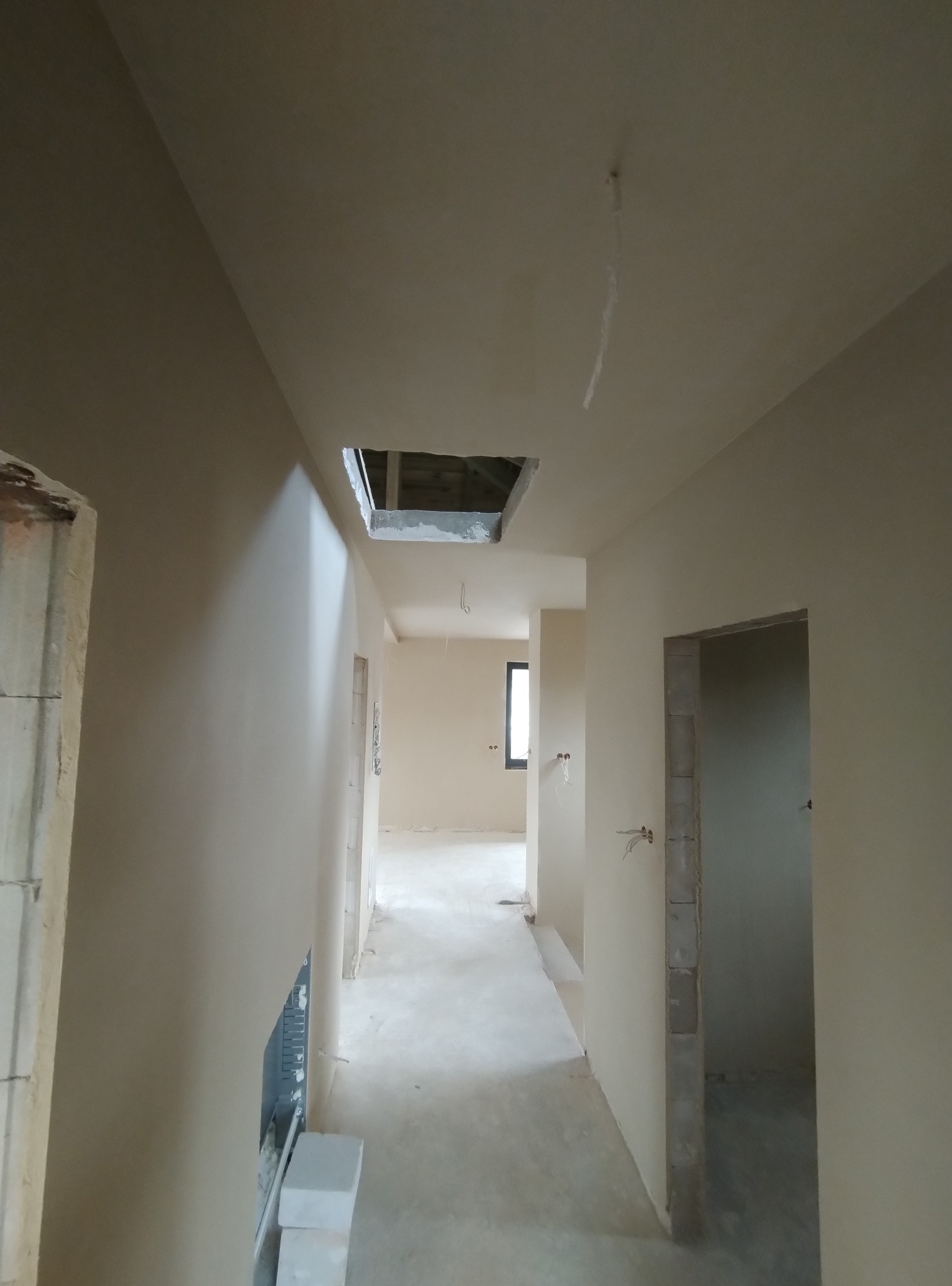 Wnętrze domu w trakcie prac wykończeniowych, widoczne świeżo nałożone, gładkie tynki na ścianach i suficie, z otworem rewizyjnym w suficie oraz wystającymi przewodami elektrycznymi.