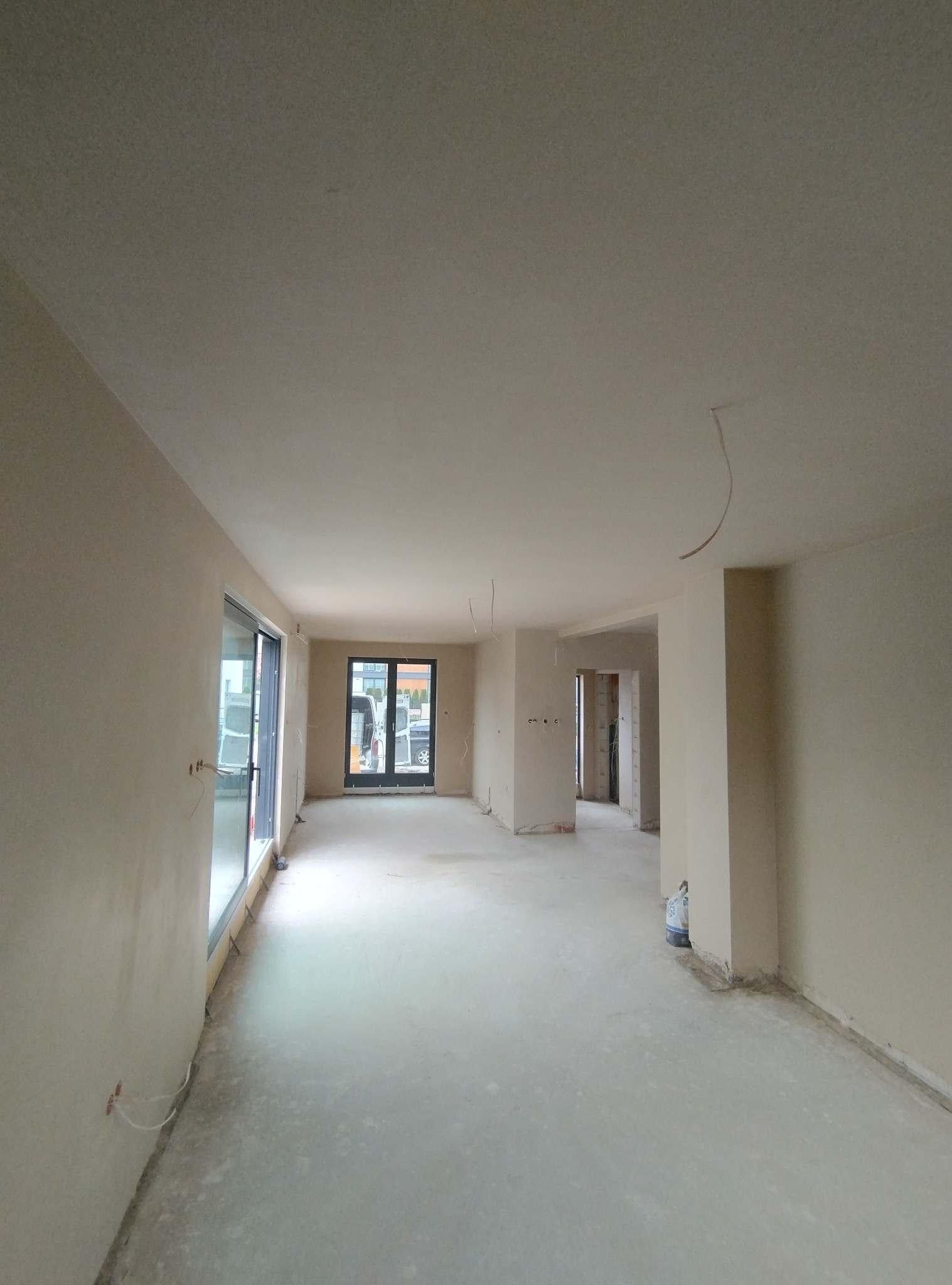 Wnętrze w stanie deweloperskim po nałożeniu gładzi, widoczne wystające kable elektryczne, okno przesuwne i drzwi balkonowe w tle.