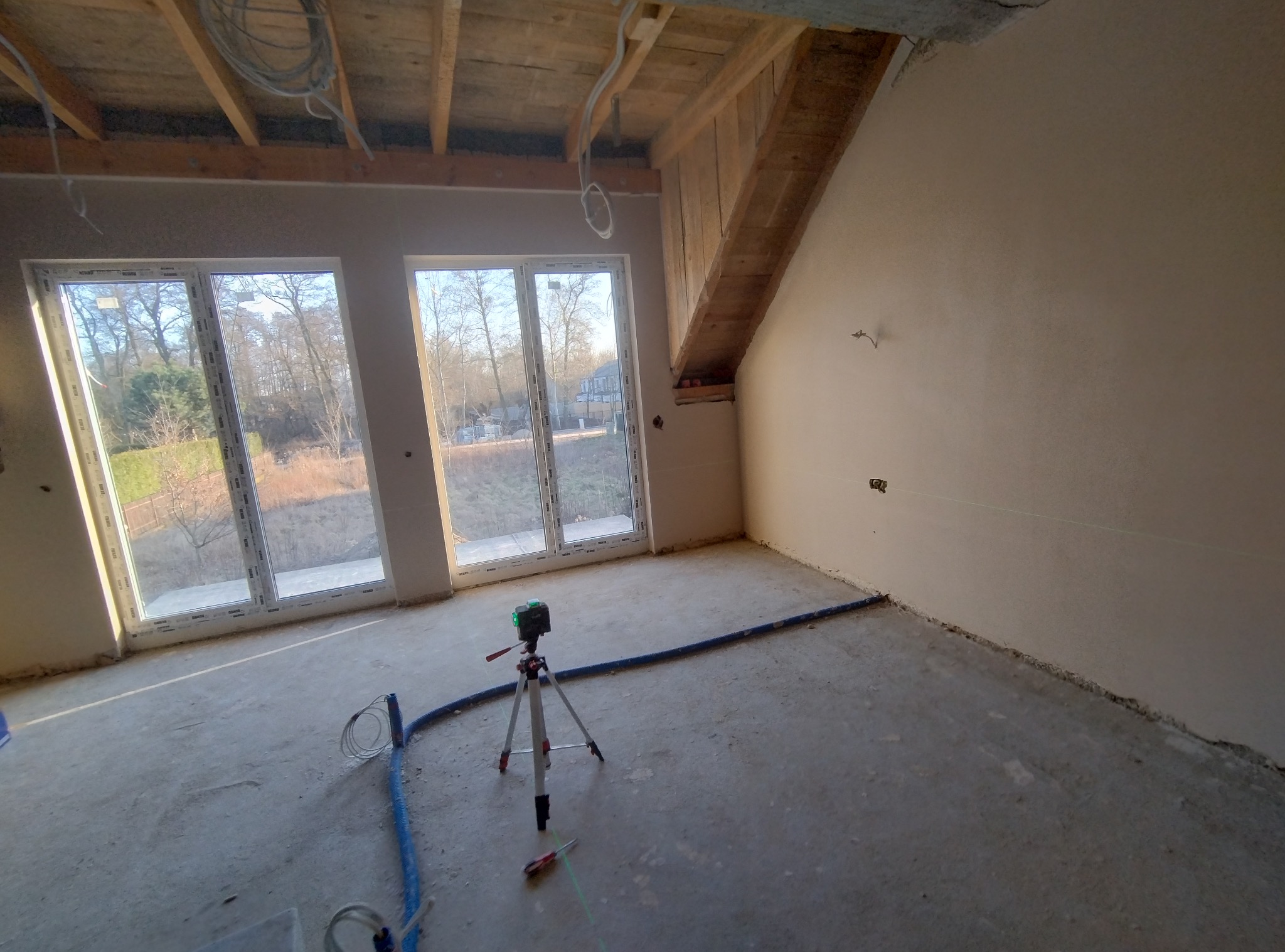 Świeżo otynkowane ściany w surowym wnętrzu domu jednorodzinnego, z widoczną drewnianą konstrukcją dachu, dużymi oknami i zielonym laserem krzyżowym na statywie wyznaczającym poziom.