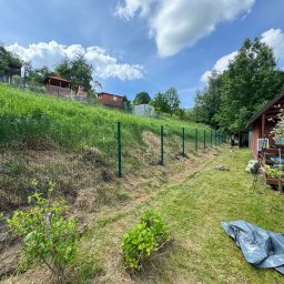 INSTAL-TUR MARCIN TURZYNIECKI - Zielone ogrodzenie panelowe na nierównym terenie z widocznymi budynkami gospodarczymi na wzgórzu, częściowo skoszona trawa i rozłożona plandeka w tle.