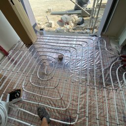 INSTAL-TUR MARCIN TURZYNIECKI - Montaż rur ogrzewania podłogowego na folii izolacyjnej z siatką, widoczne złącza i elementy instalacji, w tle okno z widokiem na pole i budowlane materiały.