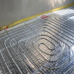 Białe rury ogrzewania podłogowego ułożone spiralnie na czarnej siatce, zabezpieczone folią, z żółtą taśmą przy ścianie, w surowym stanie wykończenia.