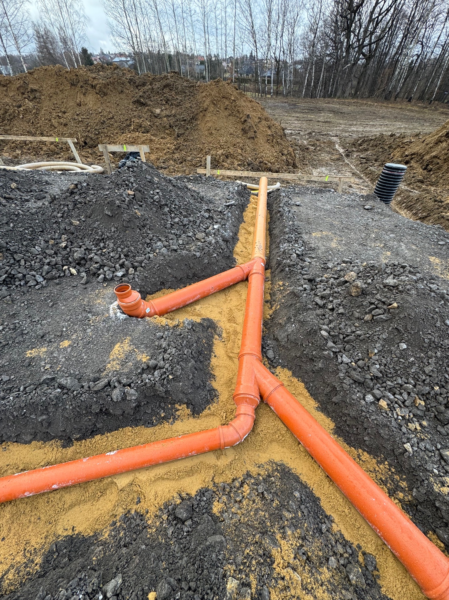Pomarańczowe rury kanalizacyjne ułożone w wykopie, podsypane piaskiem, widoczne połączenia i kształtki.