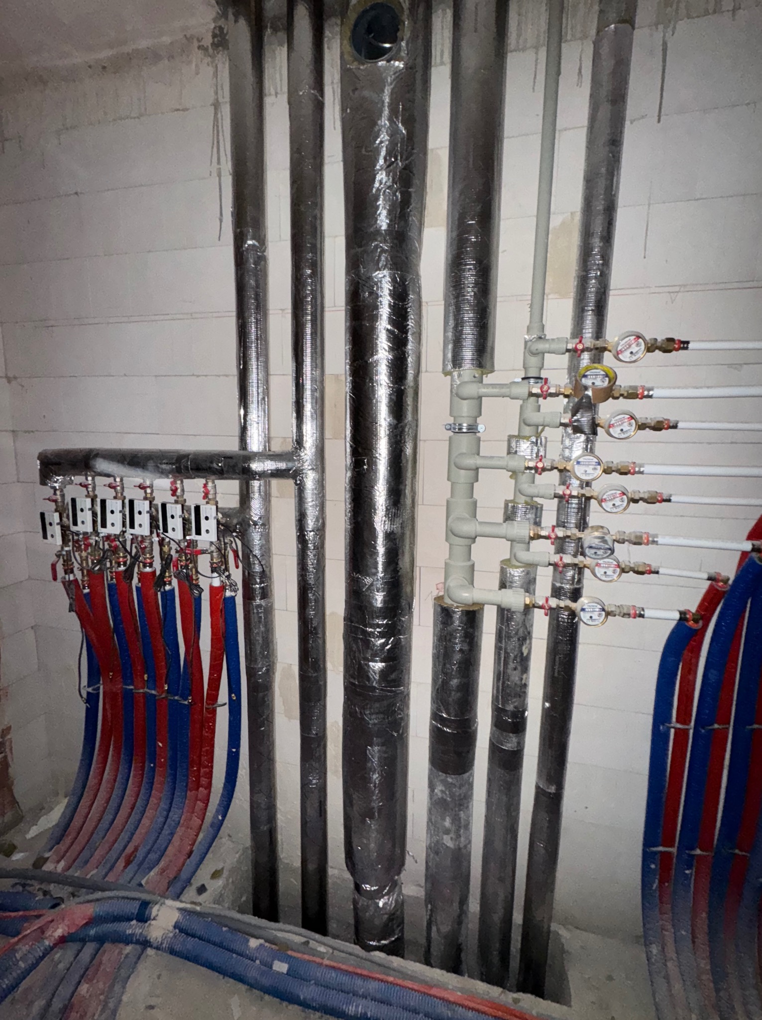 Rozdzielacz ogrzewania podłogowego z rurami PEX w kolorach czerwonym i niebieskim, obok pionowych rur owiniętych folią izolacyjną i instalacji z wodomierzami, na tle ściany z białych bloczków.