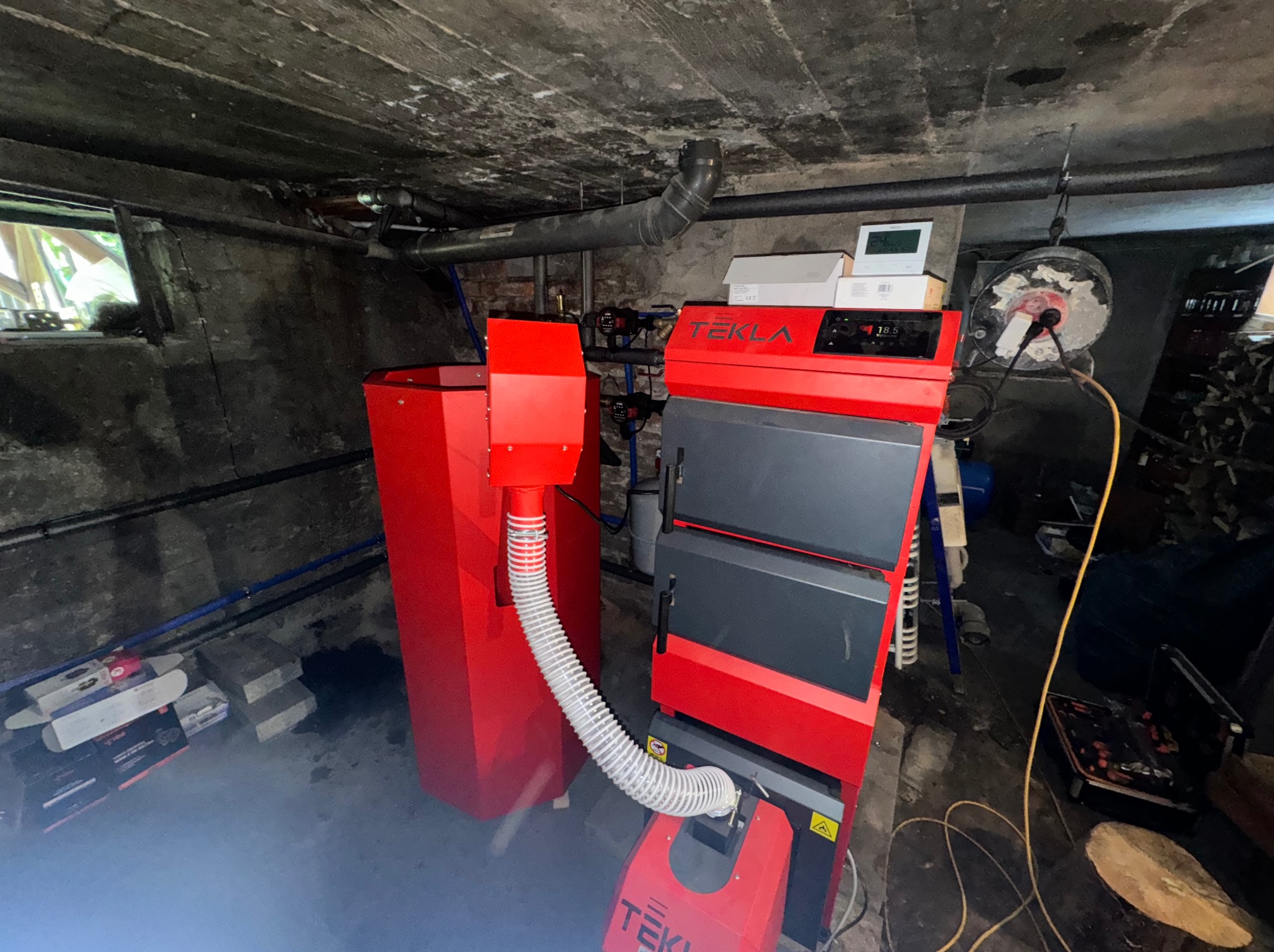 Czerwono-szary piec na pellet marki Tekla w piwnicy, z podajnikiem i rurą spiro, widoczne rury instalacji grzewczej i okno piwniczne.