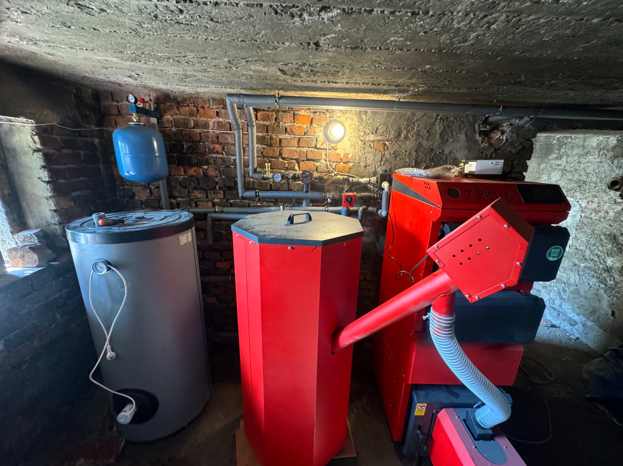 Kotłownia w Żywcu z piecem na paliwo stałe Tekla Eko, zasobnikiem ciepłej wody i zbiornikiem wyrównawczym, widoczne instalacje rurowe na tle ceglanej ściany.