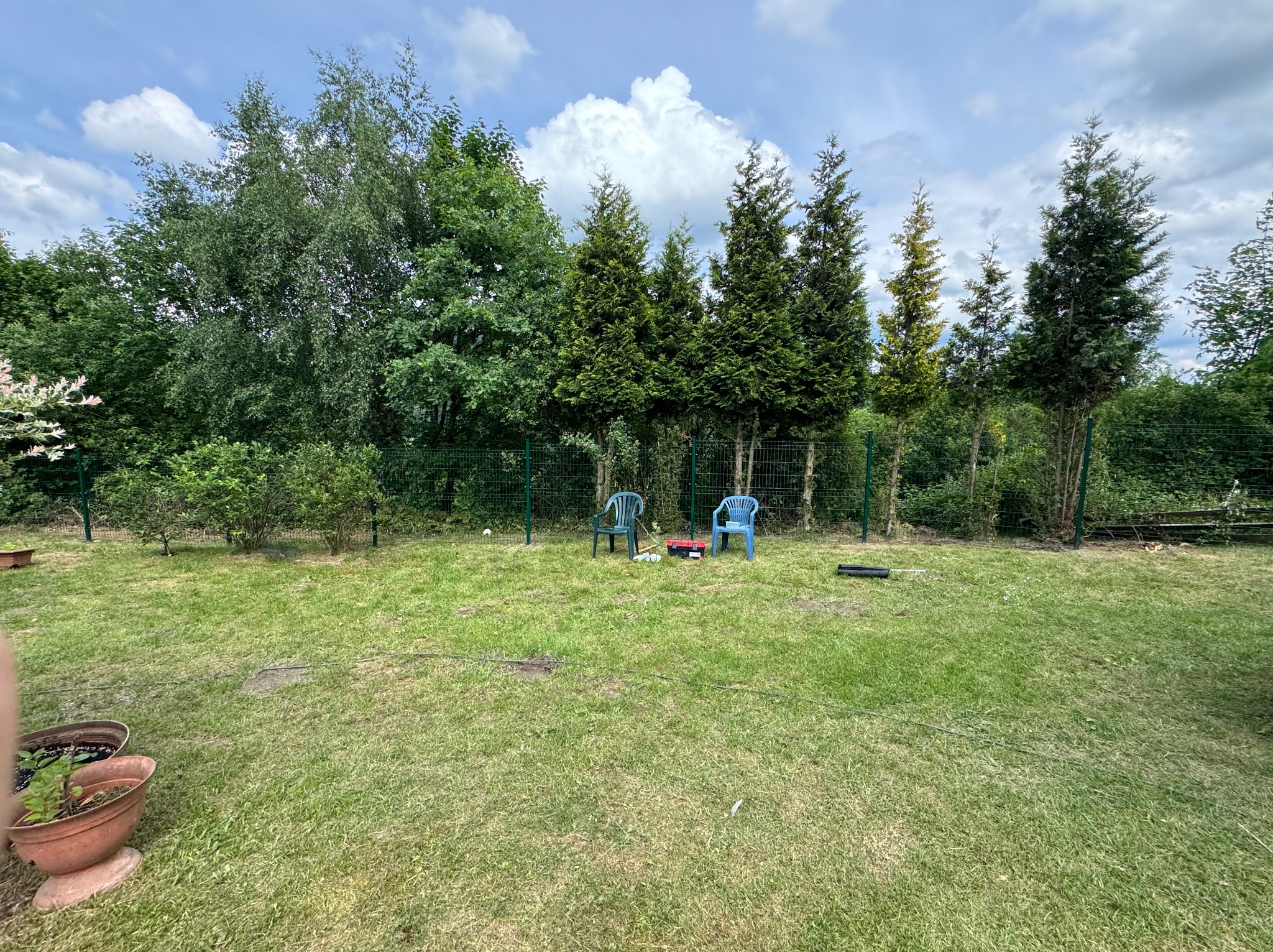 Dwa plastikowe krzesła na trawniku przed tujami, widoczne narzędzia i skrzynka narzędziowa, sugerujące prace ogrodnicze lub instalacyjne w tle zielonego ogrodzenia.