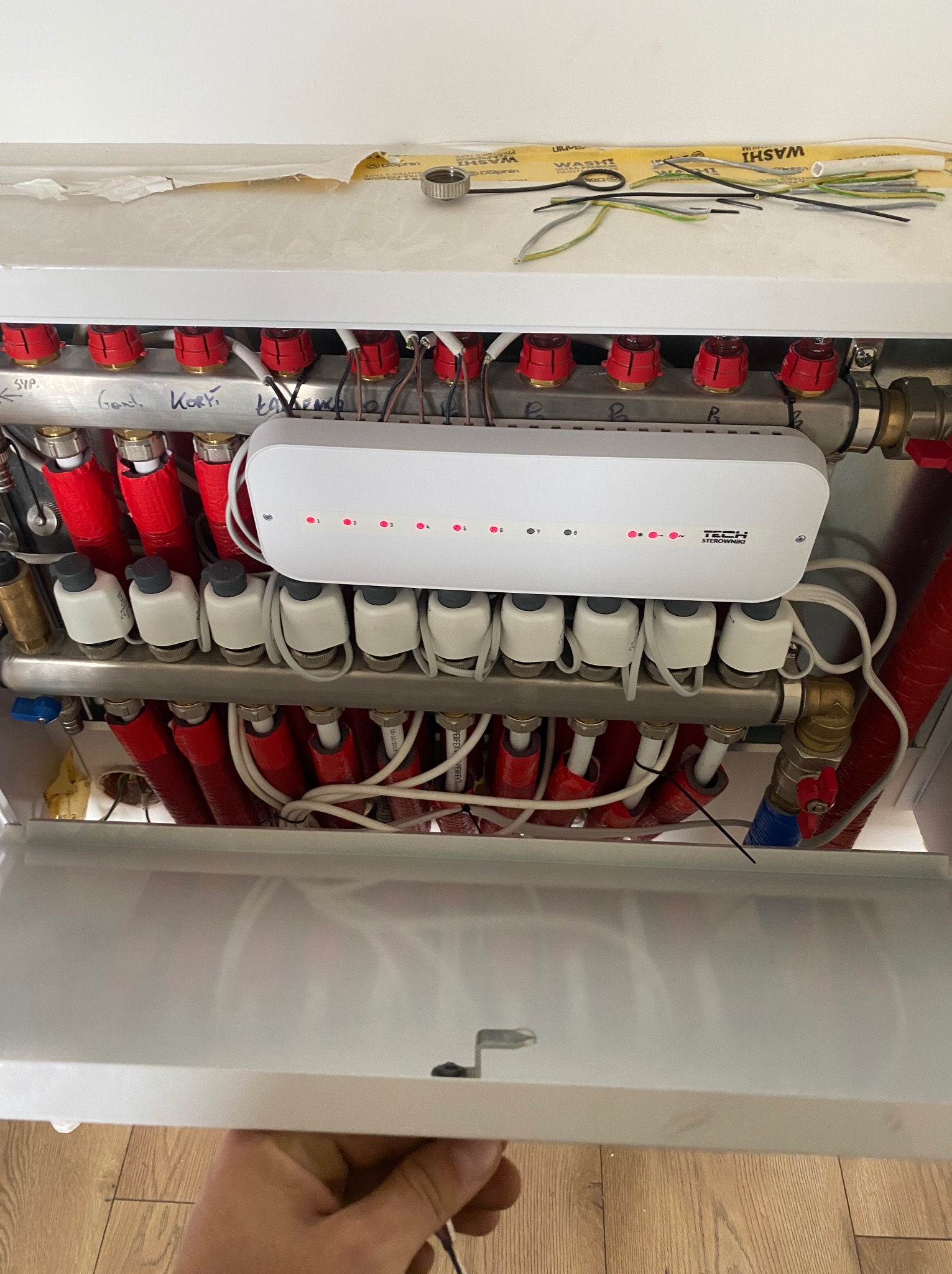 Rozdzielacz ogrzewania podłogowego z widocznym sterownikiem TECH i podłączonymi rurami w izolacji, instalacja w trakcie montażu, widoczne kable i narzędzia na górnej półce.