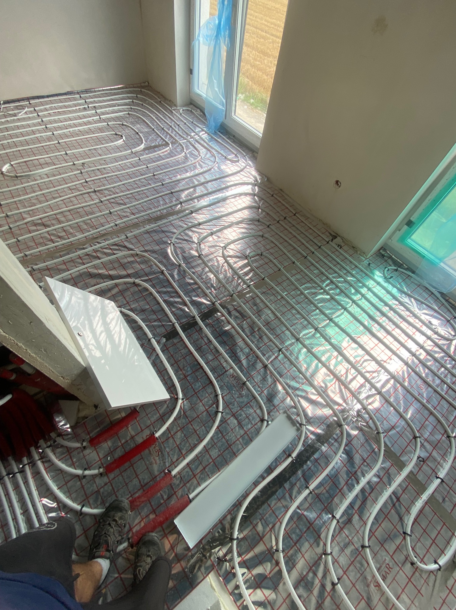 Białe rurki ogrzewania podłogowego ułożone na folii z siatką w pomieszczeniu z oknami. Widoczne buty instalatora i fragmenty płyt izolacyjnych.