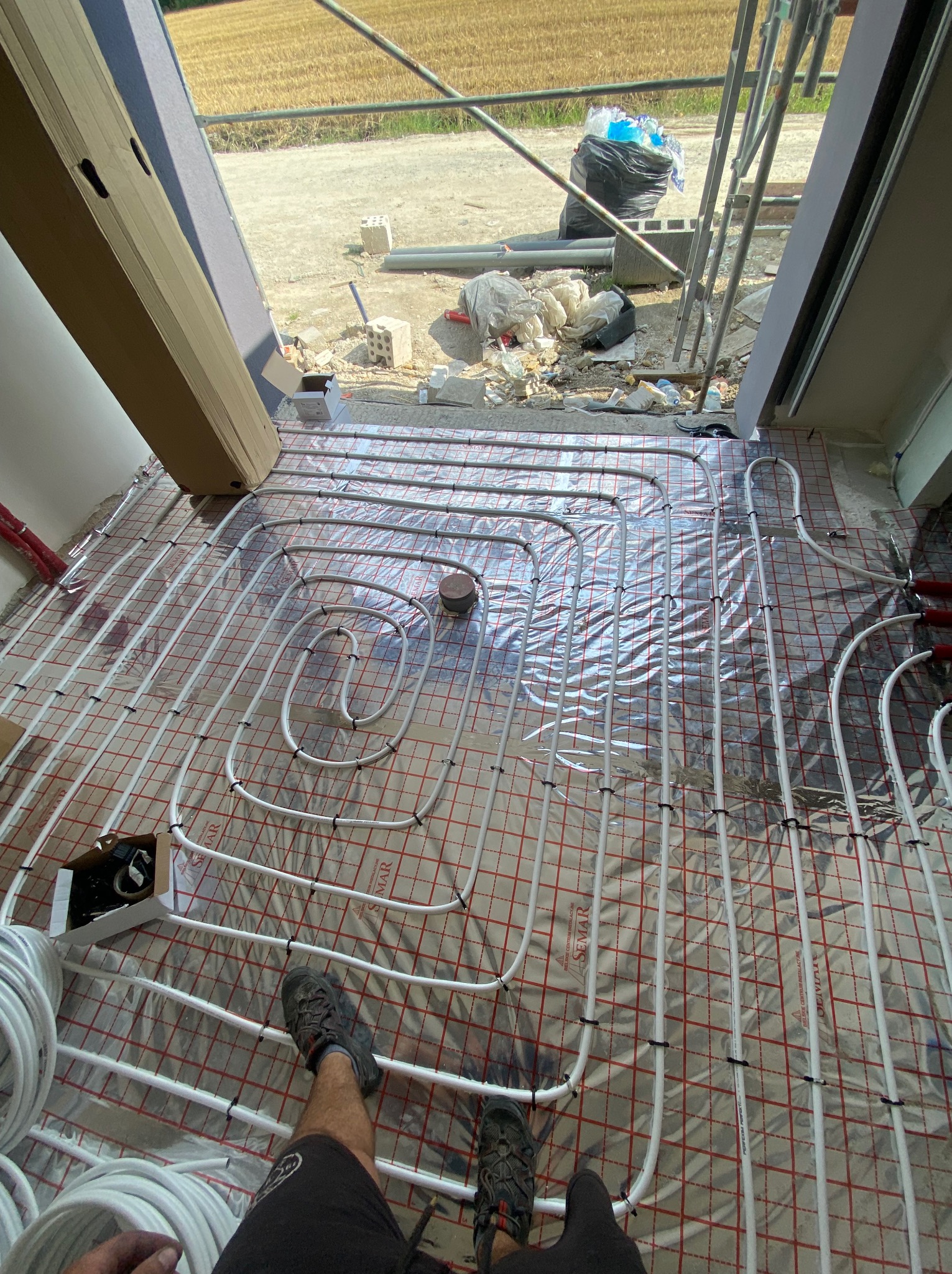 Montaż rur ogrzewania podłogowego na folii izolacyjnej z siatką, widoczne złącza i elementy instalacji, w tle okno z widokiem na pole i budowlane materiały.