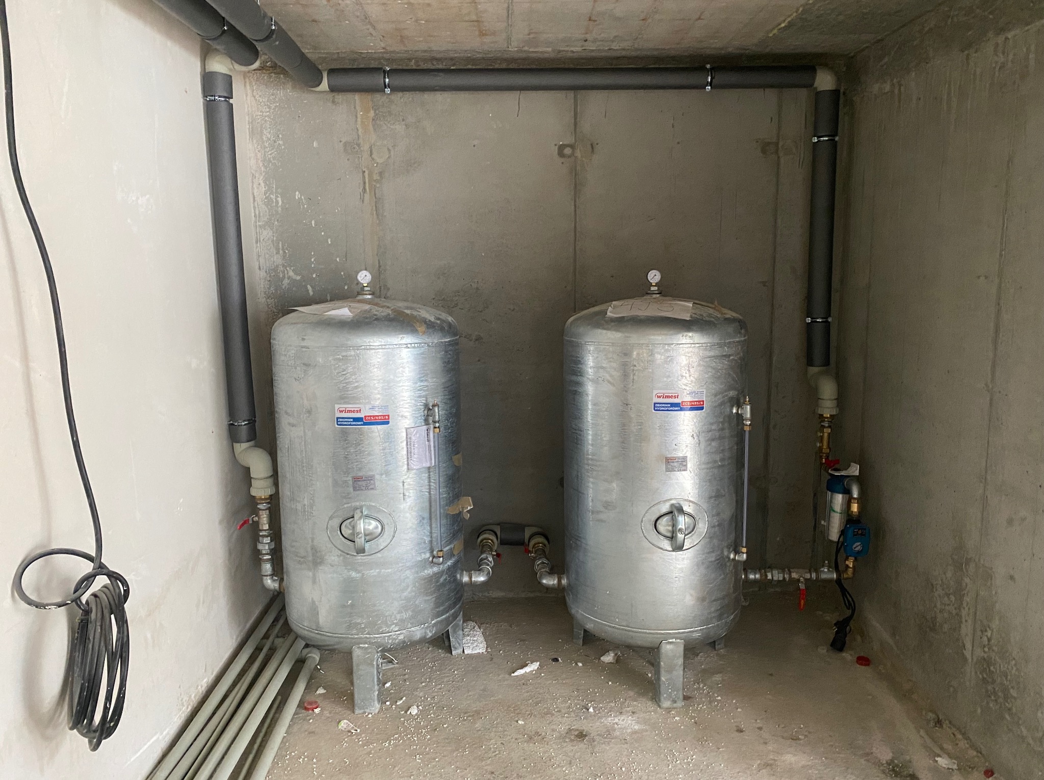 Dwa srebrne zbiorniki hydroforowe WIMEST ZCS/695/6 połączone rurami w surowym pomieszczeniu z betonowymi ścianami i sufitem, widoczne rury instalacyjne z izolacją termiczną.