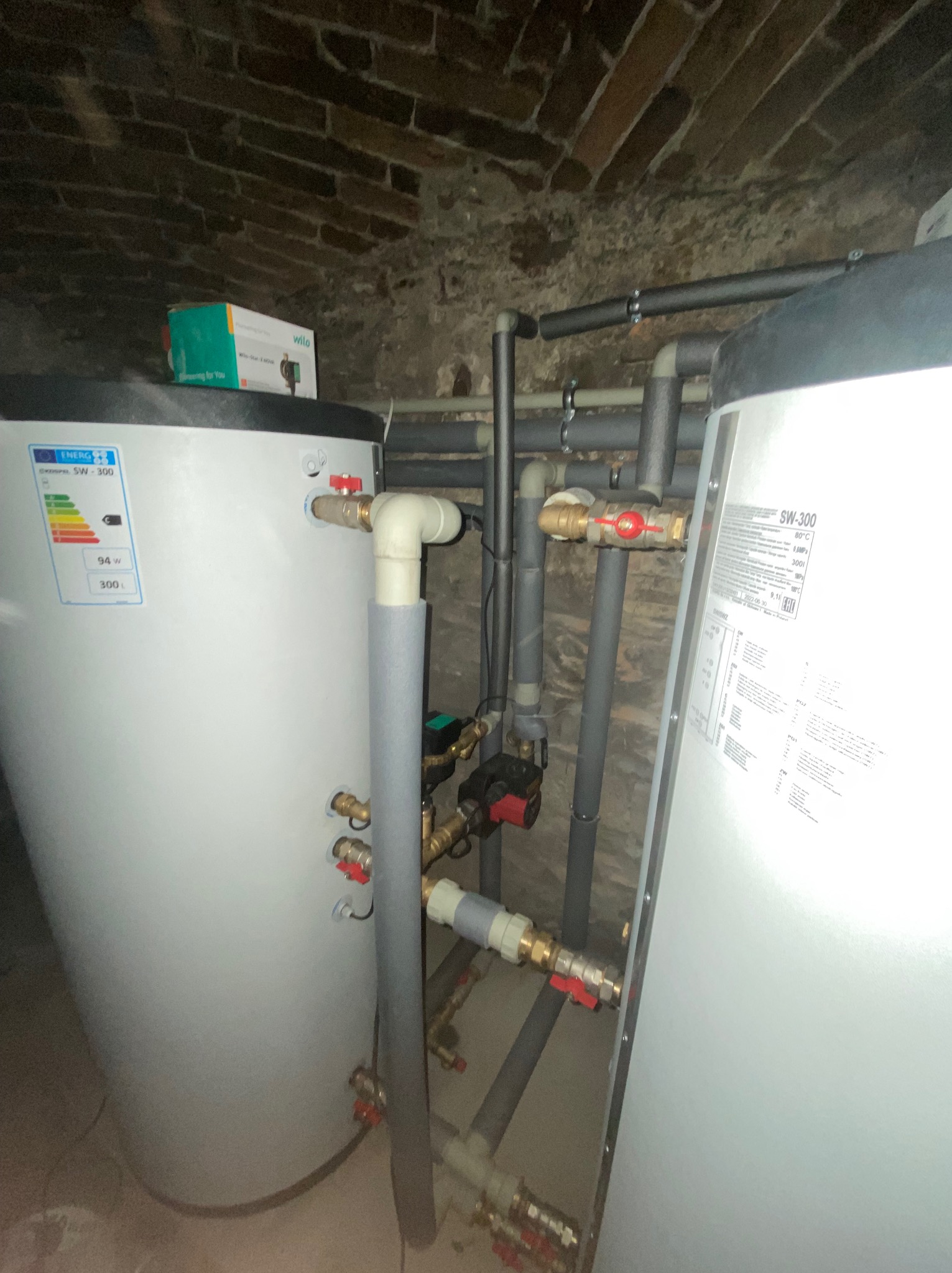 Dwa białe zasobniki ciepłej wody SW-300 w piwnicy z ceglanym sklepieniem, widoczne rury instalacyjne z zaworami, izolacja termiczna i pompa obiegowa, na jednym zbiorniku naklejka z klasą energetyczną.