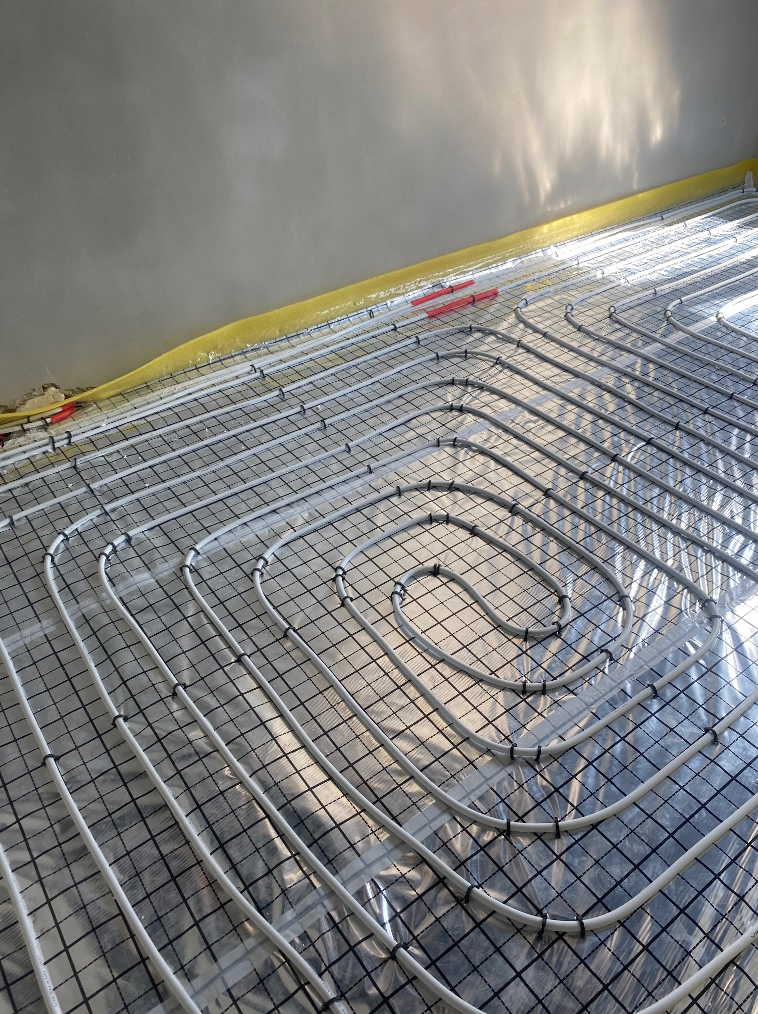 Białe rury ogrzewania podłogowego ułożone spiralnie na czarnej siatce, zabezpieczone folią, z żółtą taśmą przy ścianie, w surowym stanie wykończenia.