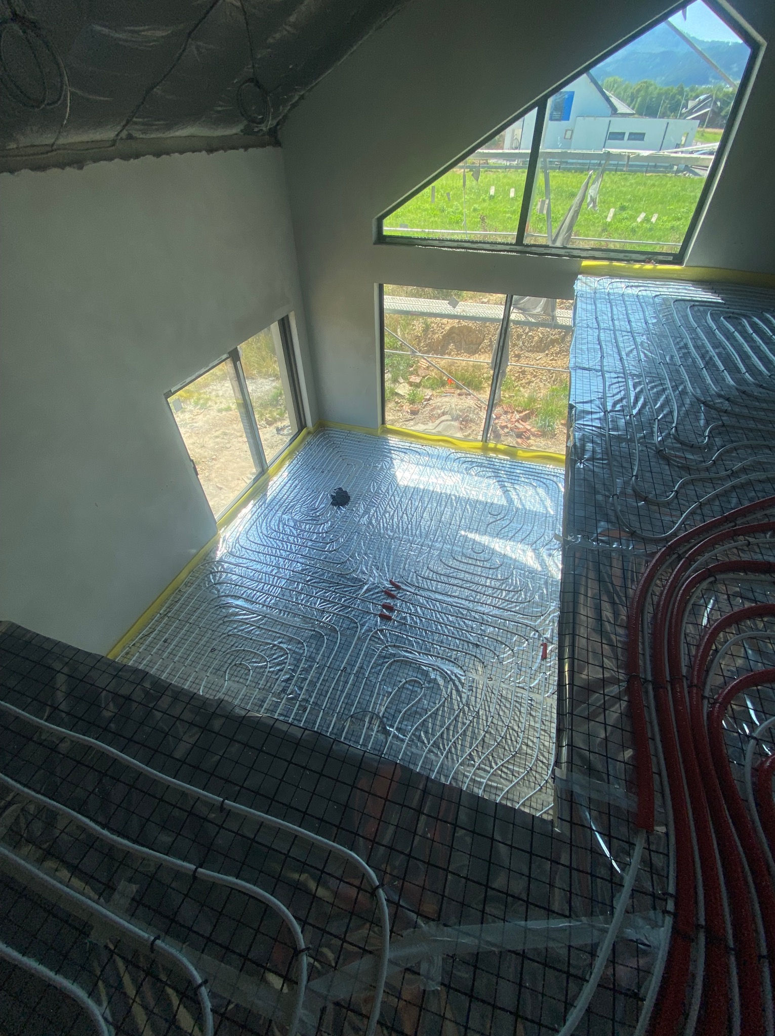 Widok z góry na rozłożoną instalację ogrzewania podłogowego w pomieszczeniu z dużymi oknami, widoczne rury grzewcze ułożone na folii izolacyjnej, przygotowane do zalania wylewką.