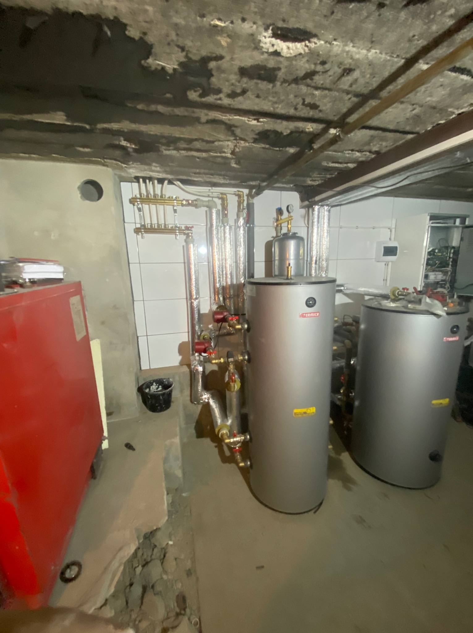 Dwa srebrne zbiorniki akumulacyjne firmy Termica w piwnicy, z widocznymi rurami i instalacją grzewczą na tle białych płytek i surowego sufitu.