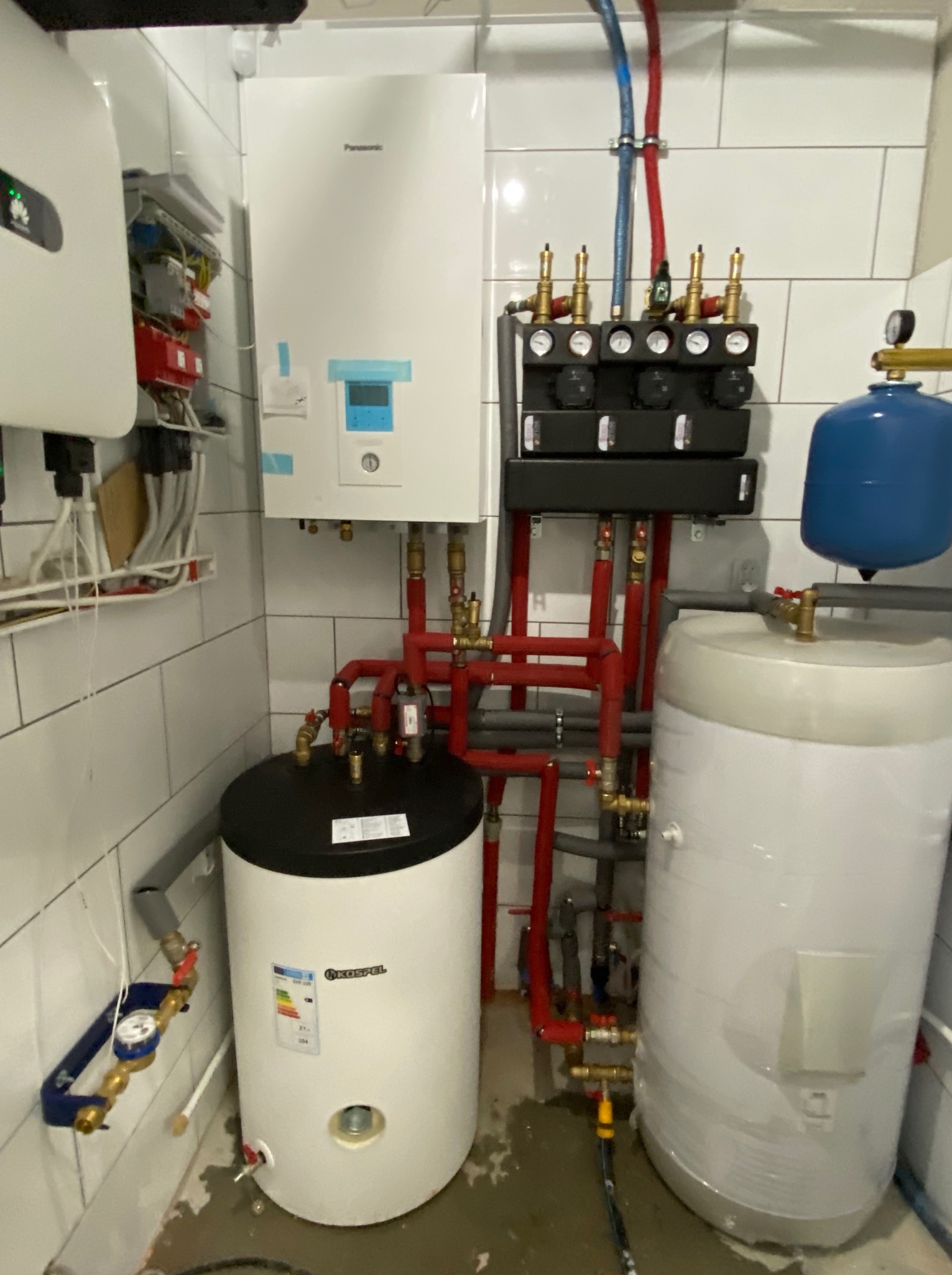 Kotłownia z białymi płytkami, zawierająca biały bojler Kospel, biały piec Panasonic, rozdzielacz z manometrami, inwerter fotowoltaiczny Huawei i czerwone rury instalacji grzewczej.