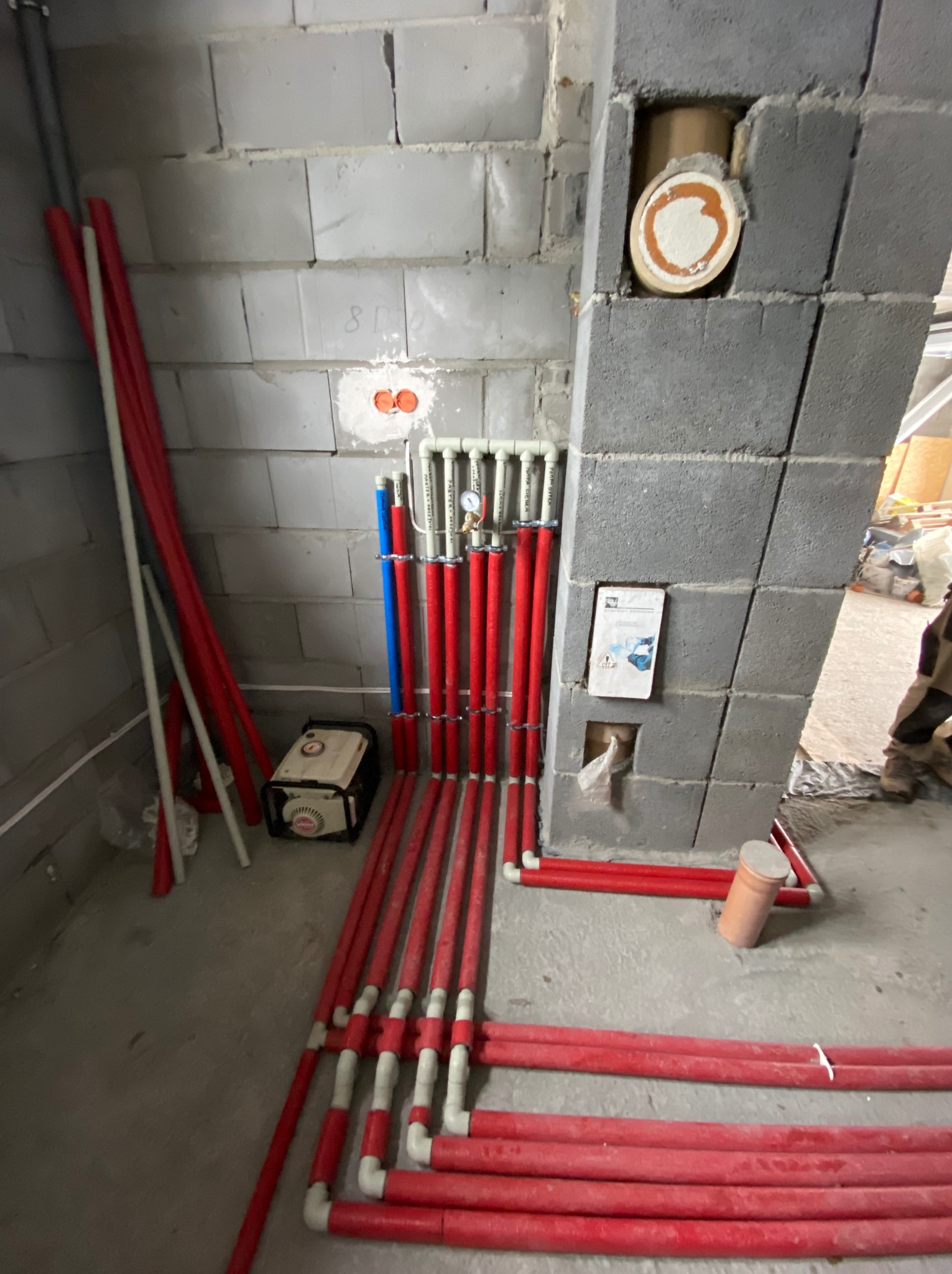 Wewnętrzne instalacje z czerwonych rur, częściowo zamontowane do ściany z betonu komórkowego, widoczny manometr i agregat prądotwórczy w tle.